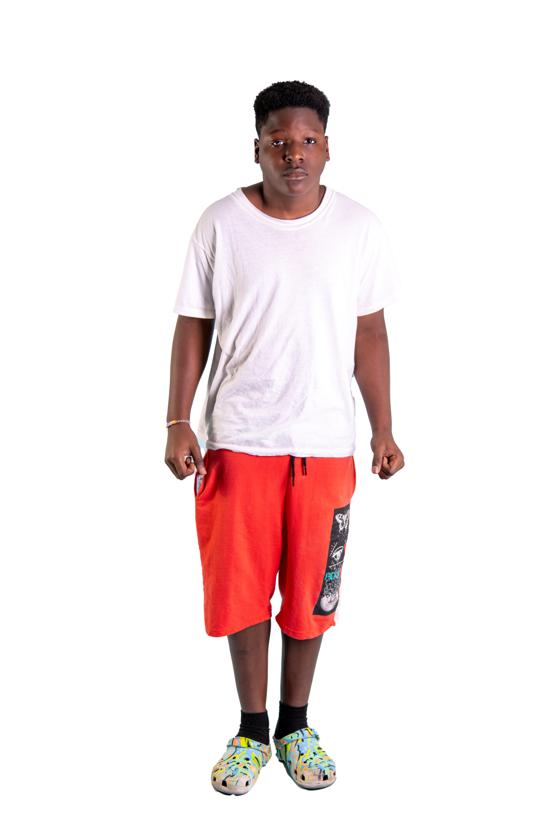 A young boy wearing a white t-shirt, red shorts, black socks, and colorful Crocs standing with his fists clenched.