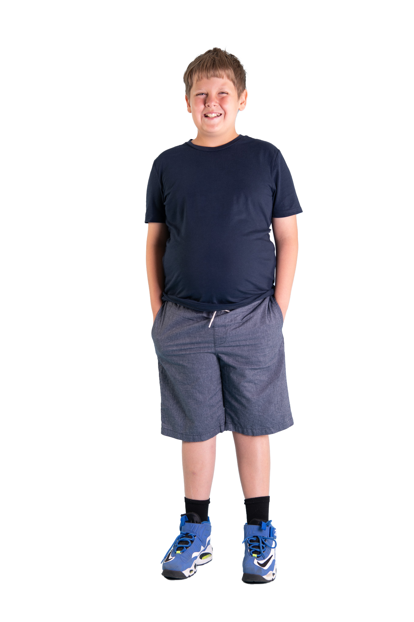 A young boy wearing a navy t-shirt, gray shorts, black socks, and blue athletic shoes, smiling with hands in pockets.