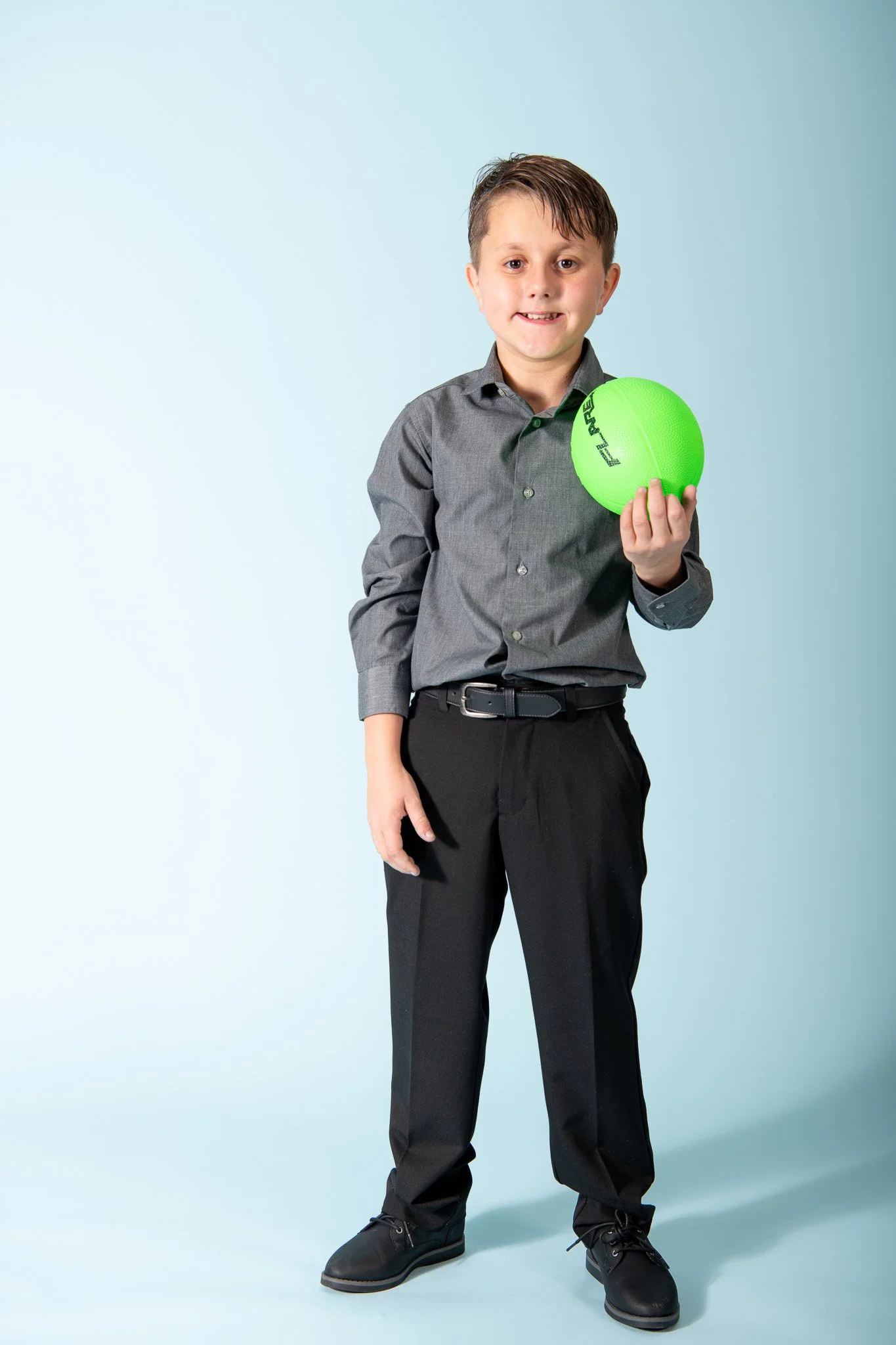 A young boy in a gray shirt and black pants holding a bright green ball.