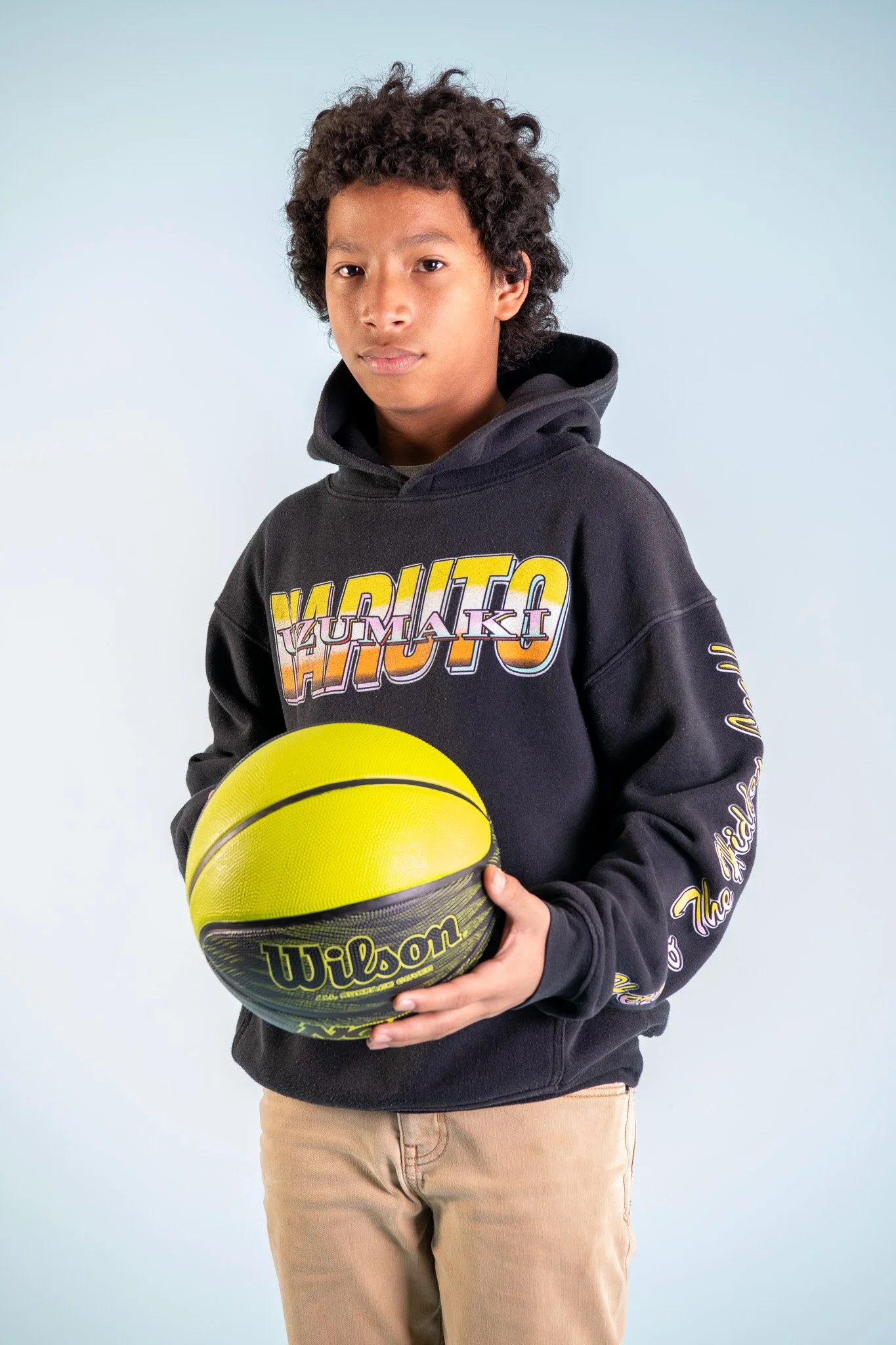 A young boy with curly hair wearing a black hoodie with colorful text and holding a yellow and black Wilson basketball.