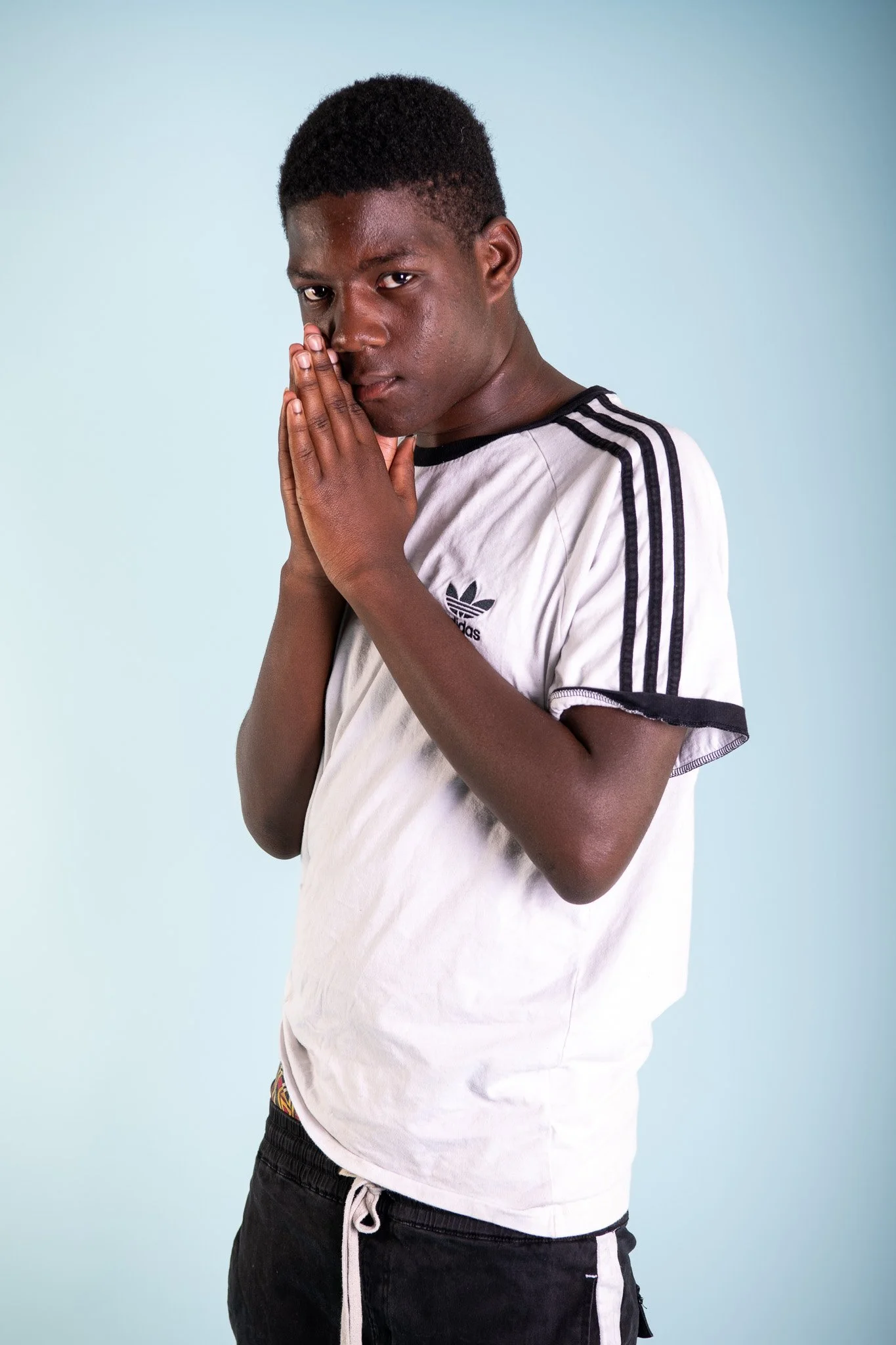 Young man with short curly hair and brown skin, standing against a light blue background, wearing a white Adidas t-shirt with black stripes and black shorts, looking at the camera with his hands pressed together near his face.
