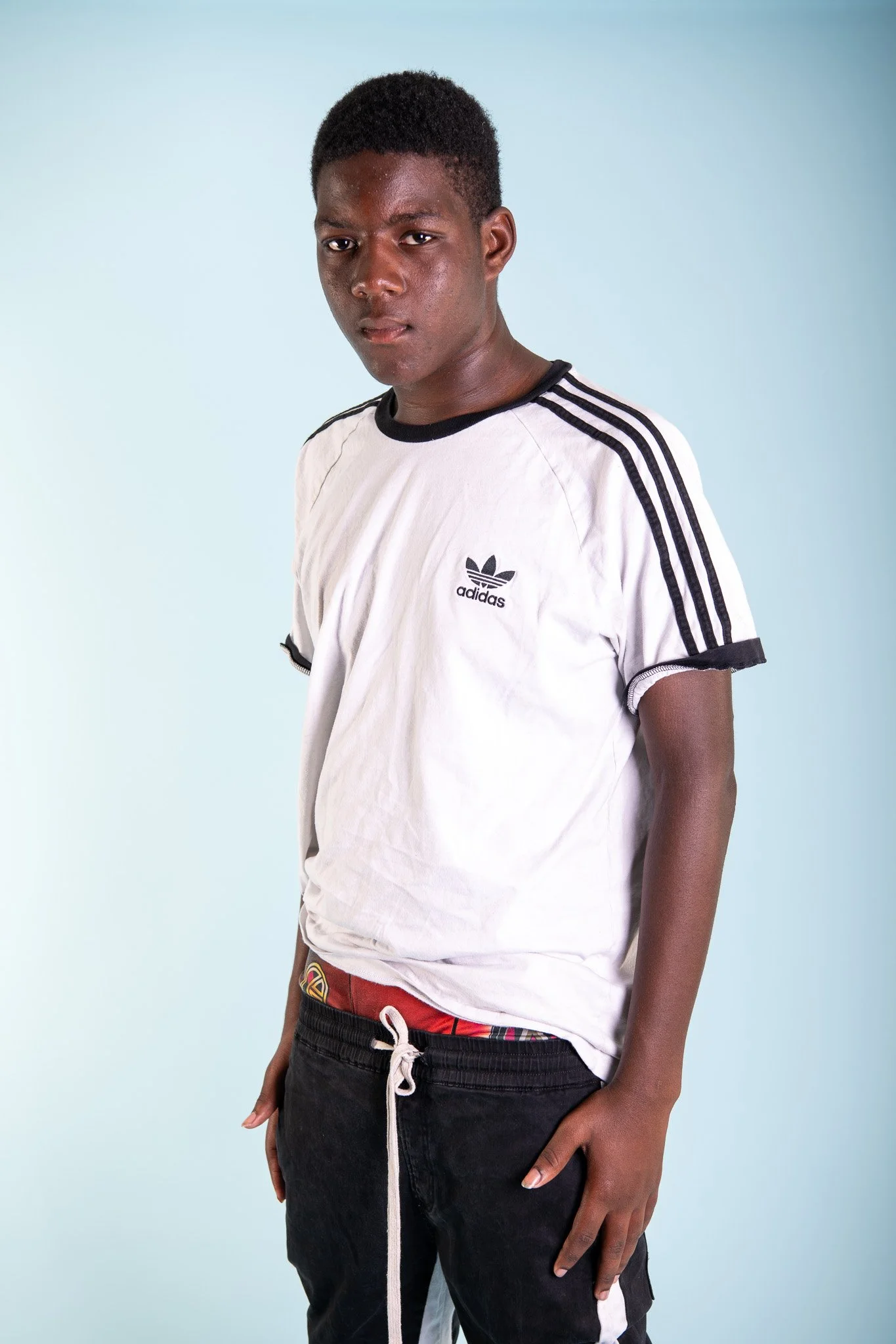 A young man with dark skin and short curly hair wearing a white Adidas t-shirt with black stripes, black pants, and red underwear, standing against a plain light blue background.