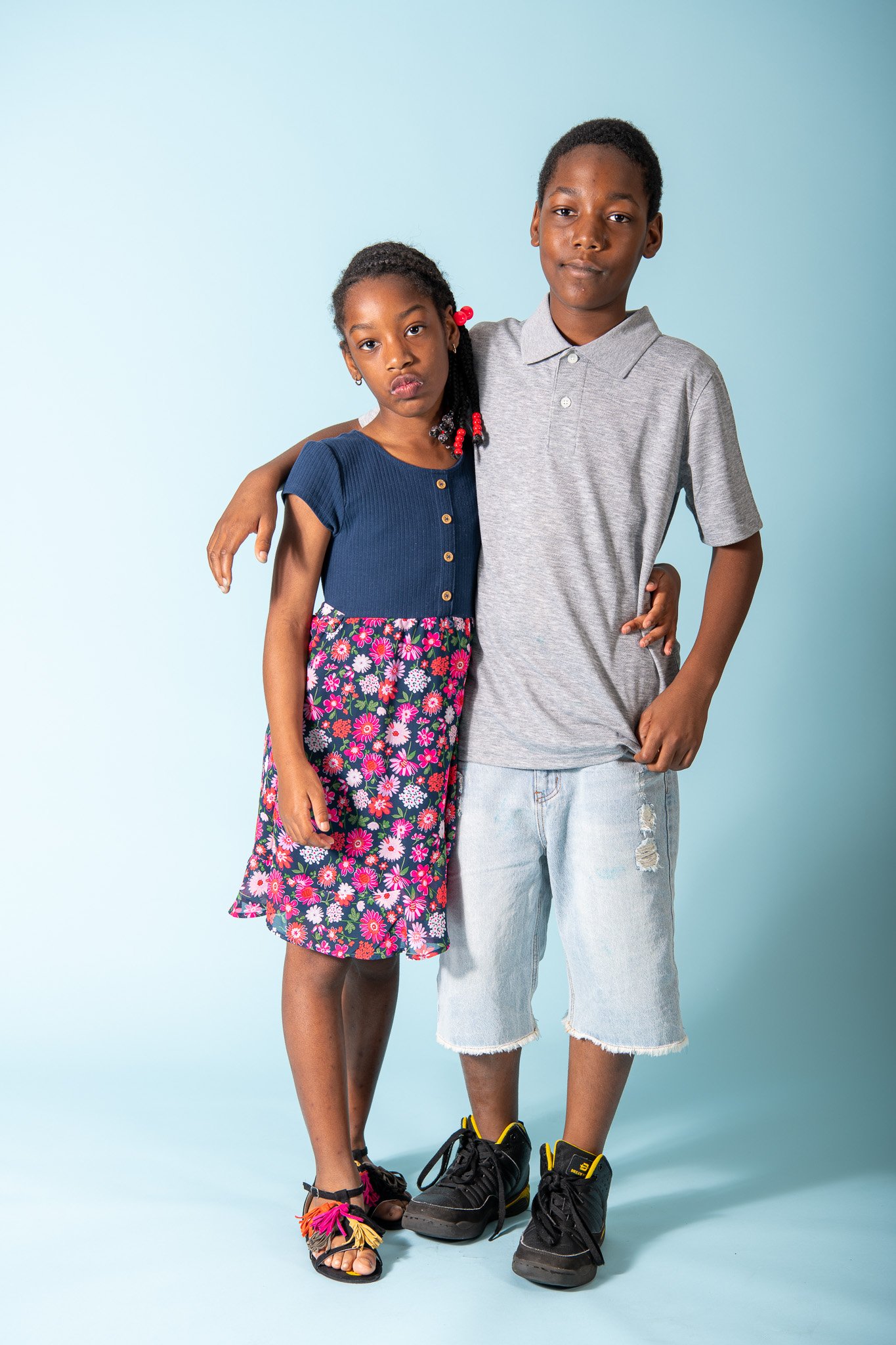 A boy and a girl stand together against a light blue background, with the boy's arm around the girl's shoulder. The girl has braided hair decorated with red beads and wears a blue top with wooden buttons and a colorful floral skirt. The boy has short