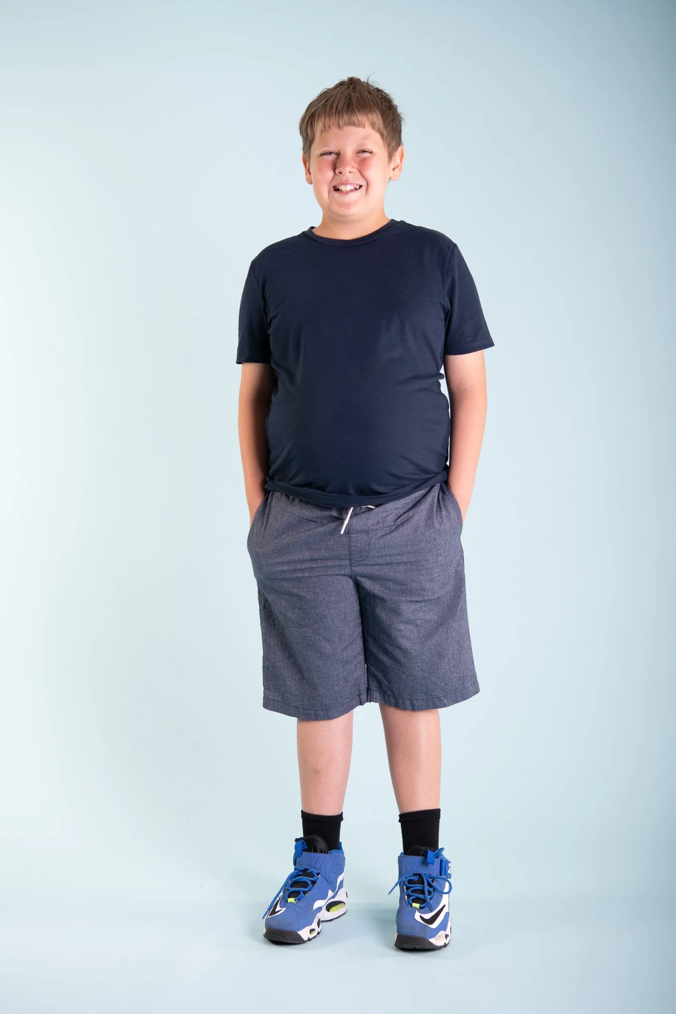 A young boy smiling, standing against a plain light-colored background, wearing a navy blue t-shirt, gray shorts, black socks, and blue sneakers.
