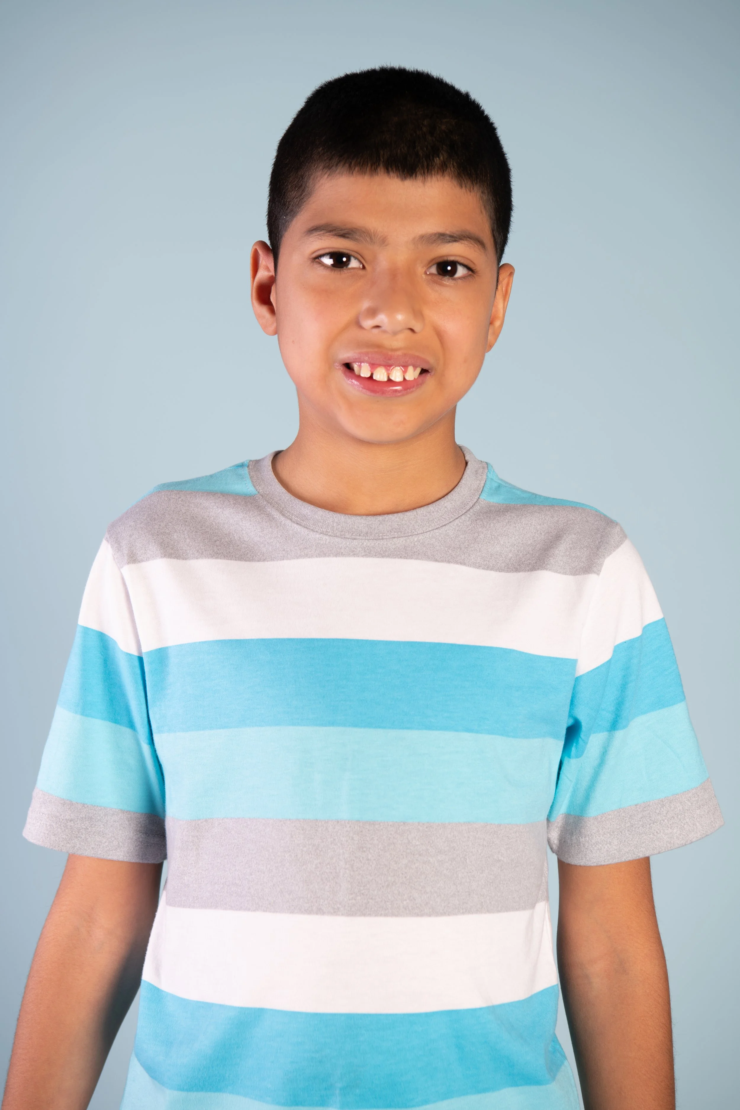 A young boy with short black hair, wearing a gray, white, and sky blue striped t-shirt, smiling in front of a light blue background.