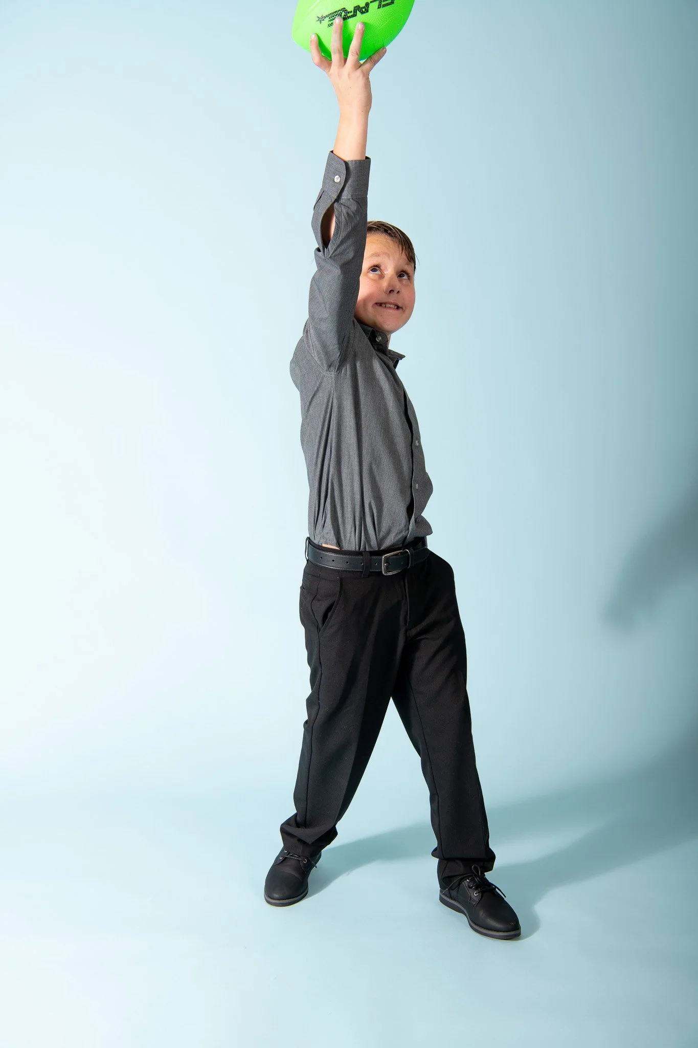 A young boy in a gray dress shirt and black pants holding a green frisbee high above his head.