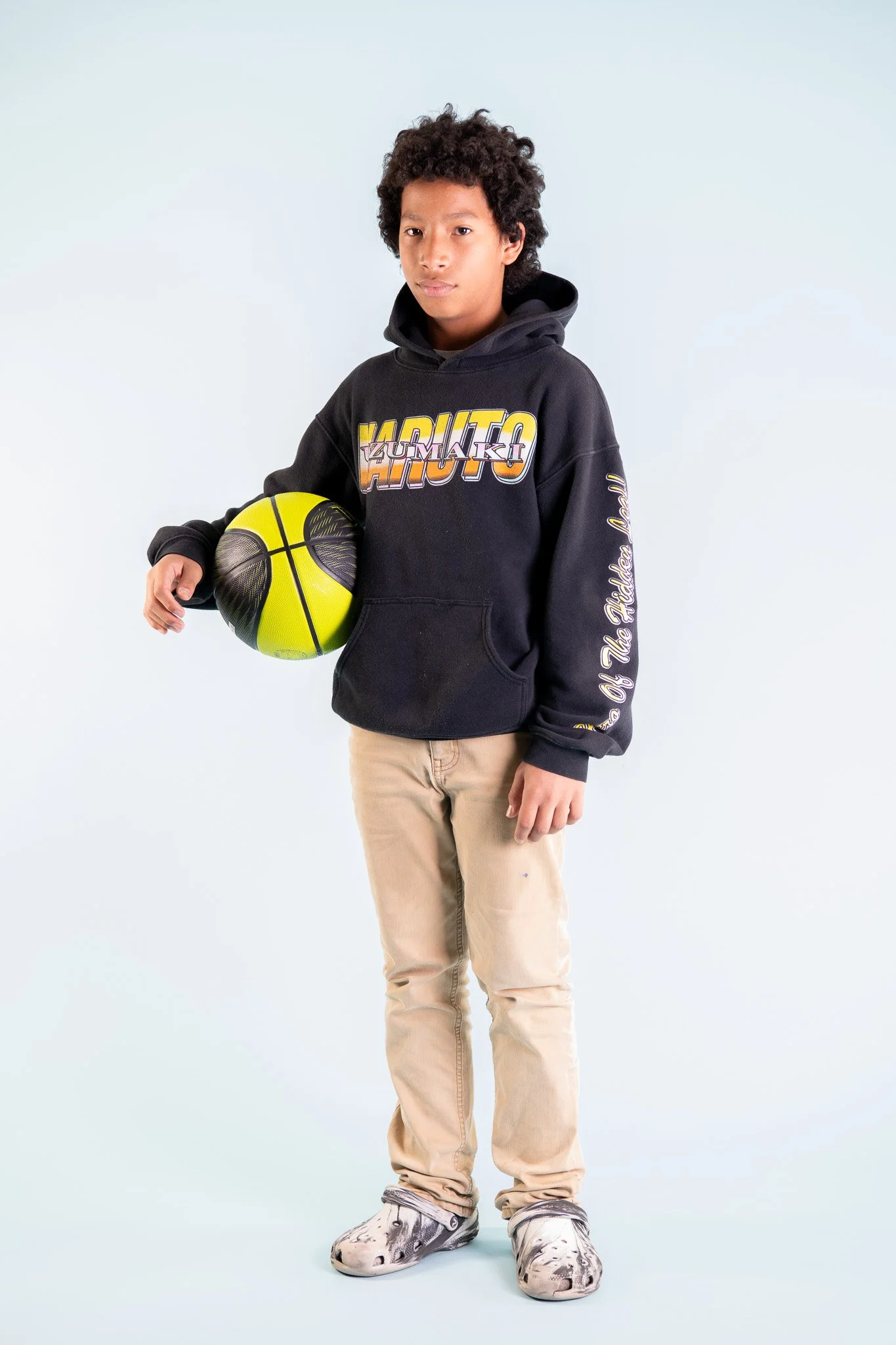 A young boy with curly hair holding a basketball, wearing a black hoodie with colorful text, beige pants, and spotted sneakers, standing against a plain white background.