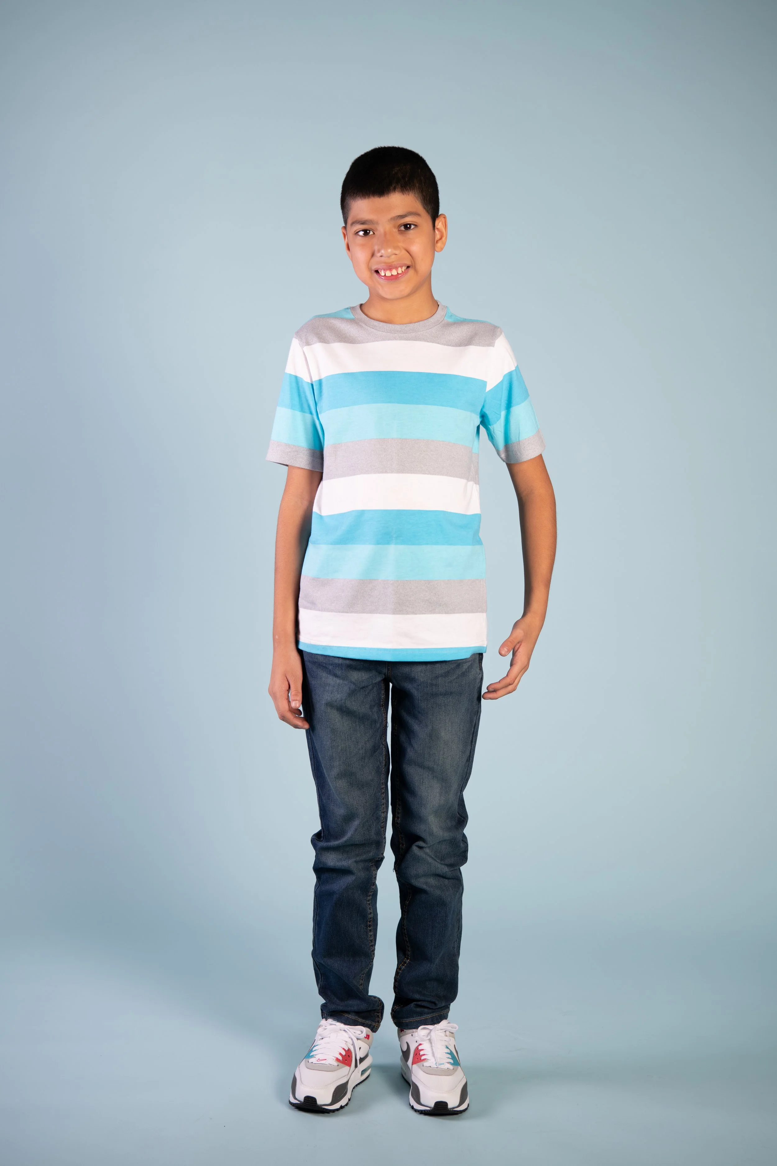 A young boy wearing a striped t-shirt, jeans, and sneakers standing against a plain light blue background.