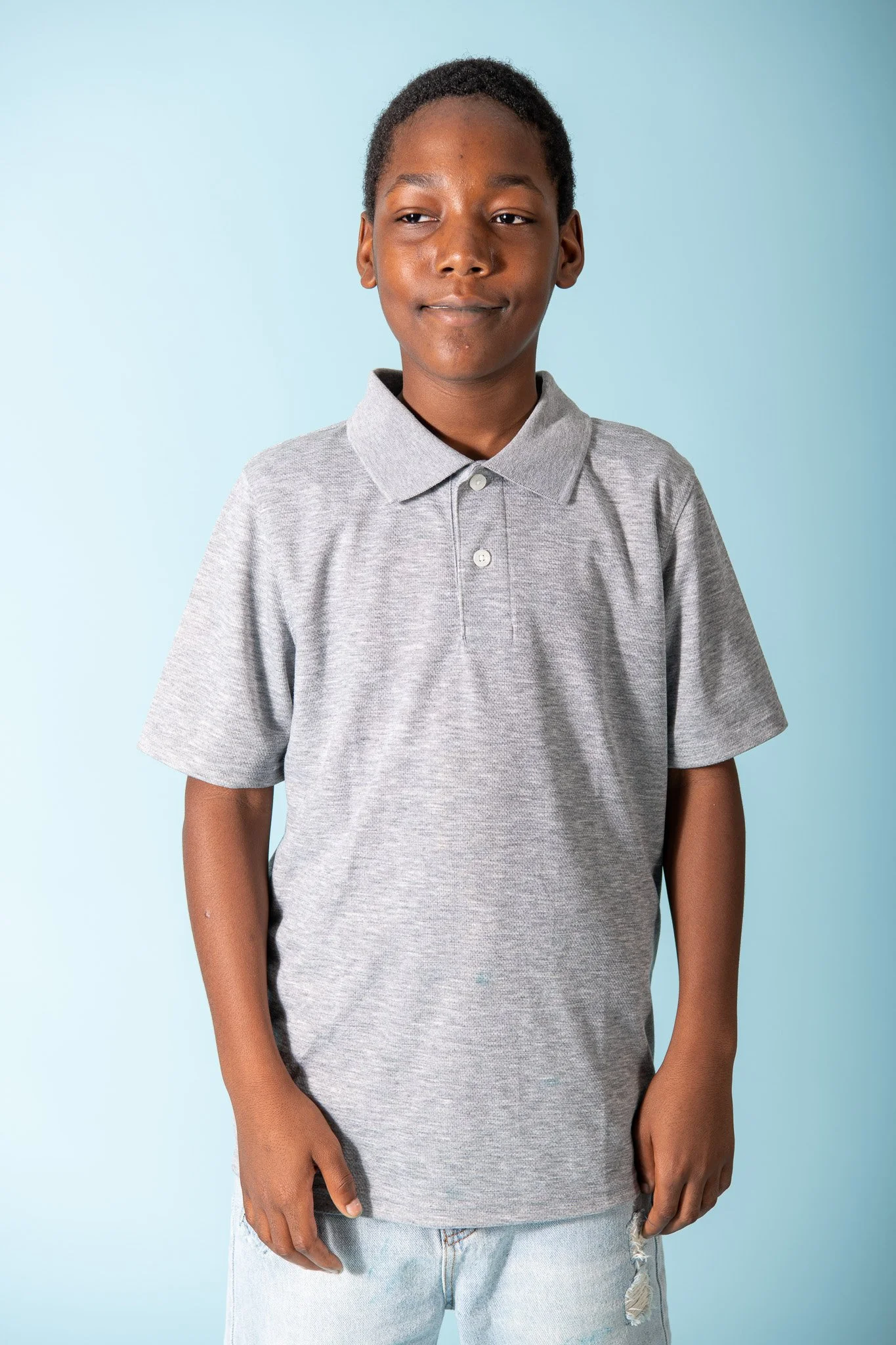 A young boy with short curly hair wearing a light gray polo shirt and light blue ripped jeans, standing against a light blue background.