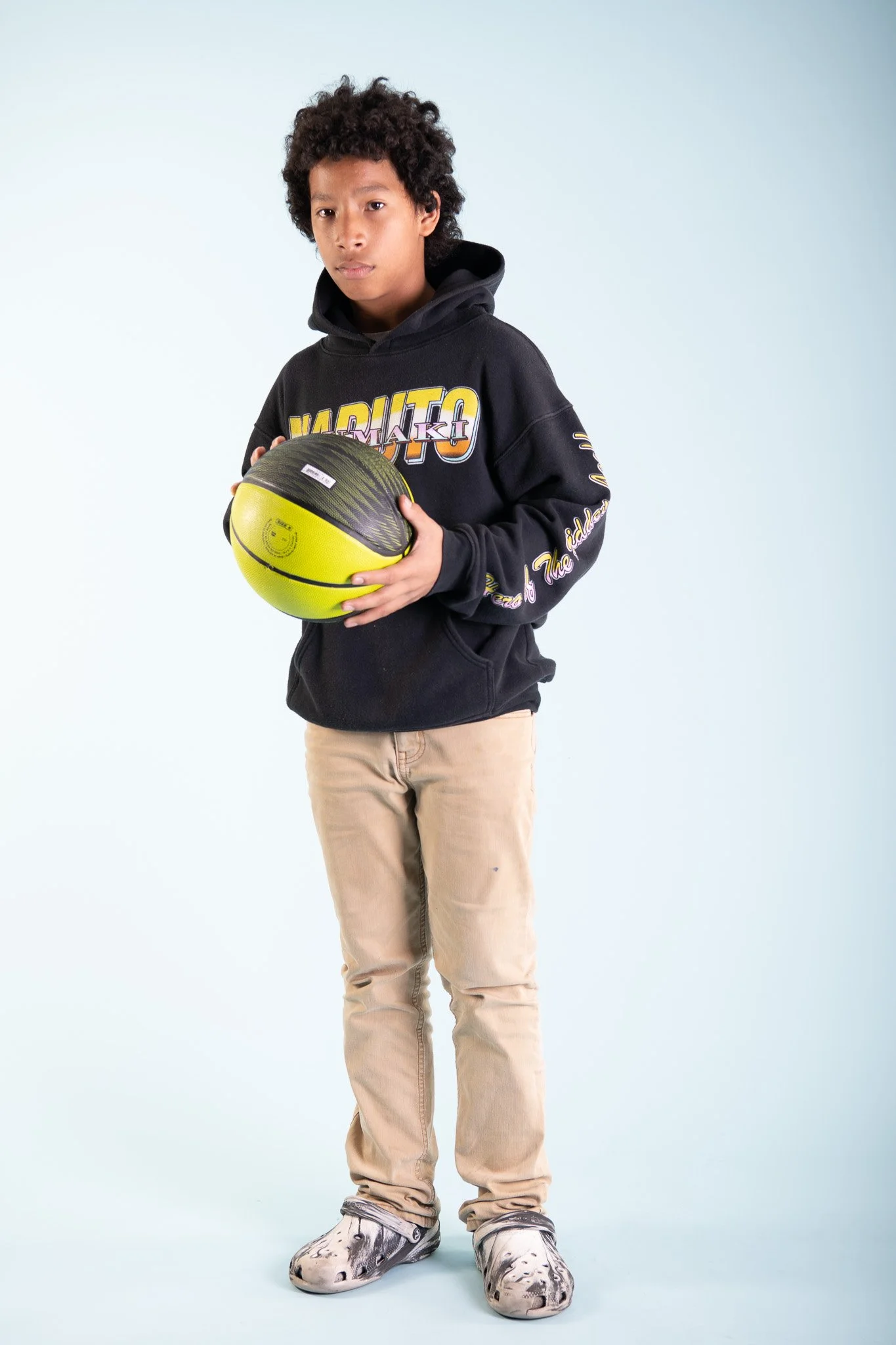 A young person standing against a light blue background holding a yellow and black basketball.