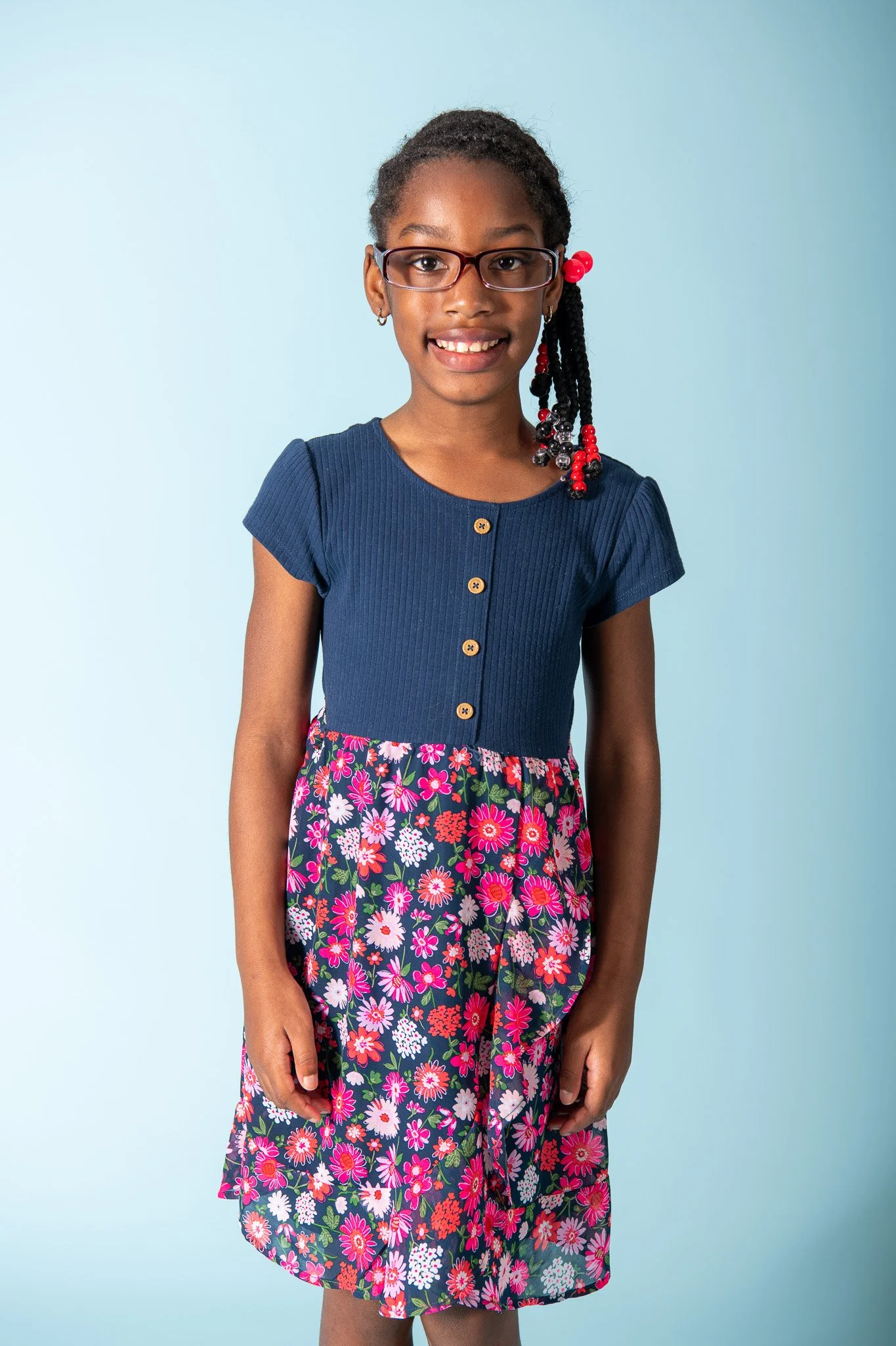 Young girl smiling, wearing glasses with red beads in braided hair, blue top, and floral skirt, against a light blue background.