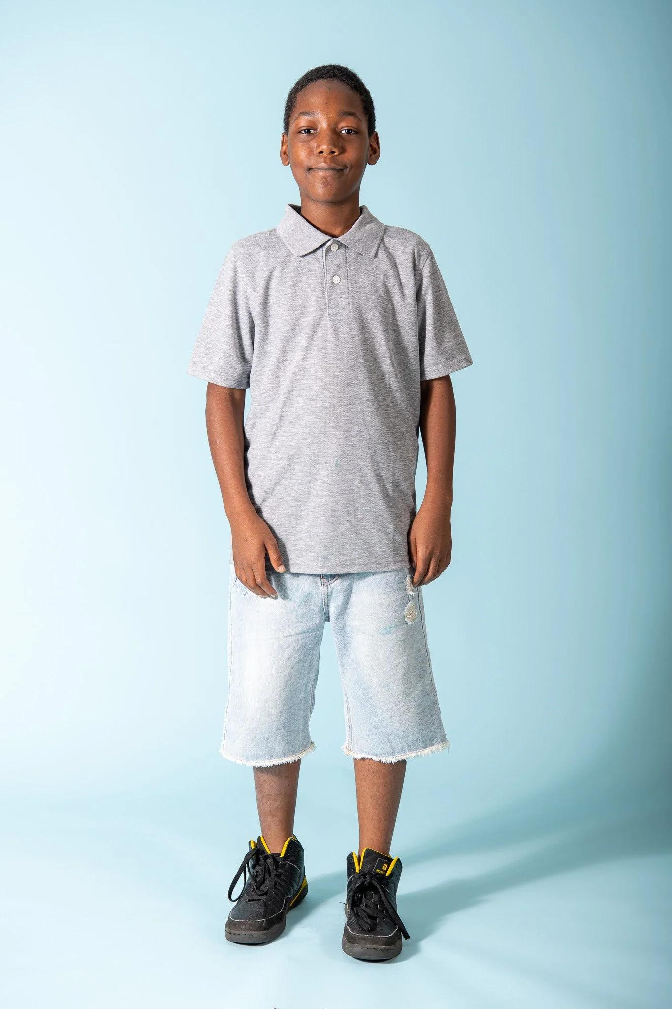 A young boy standing against a light blue background, wearing a gray polo shirt, distressed denim shorts, and black sneakers with yellow accents.