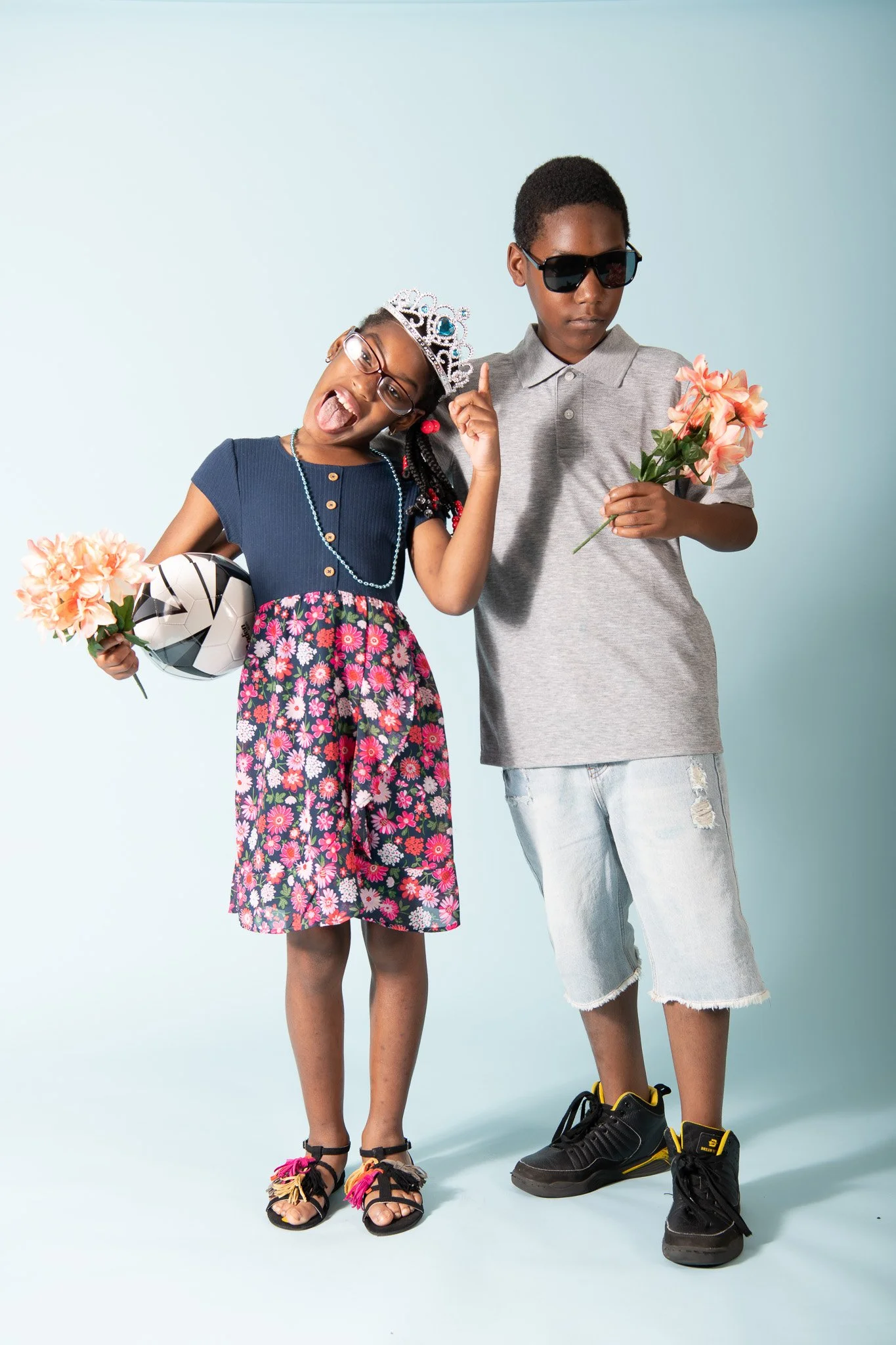 A girl wearing glasses, a tiara, and a blue top with a floral skirt, holding a flower and a soccer ball, making a playful expression. A boy in a gray shirt and ripped shorts, wearing sunglasses, holding a flower, standing beside her against a light b