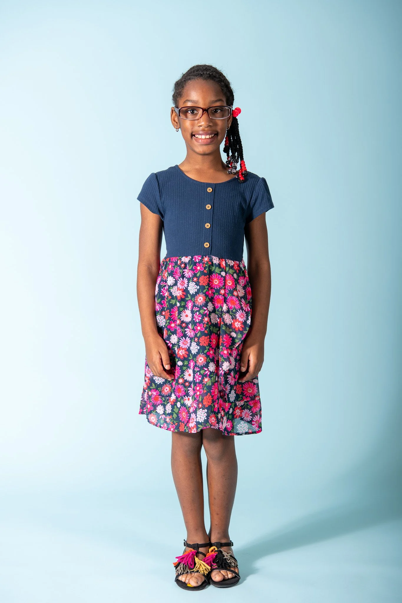 A young girl with glasses and braids, smiling, wearing a navy blue shirt with gold buttons and a floral skirt, standing on a light blue background.