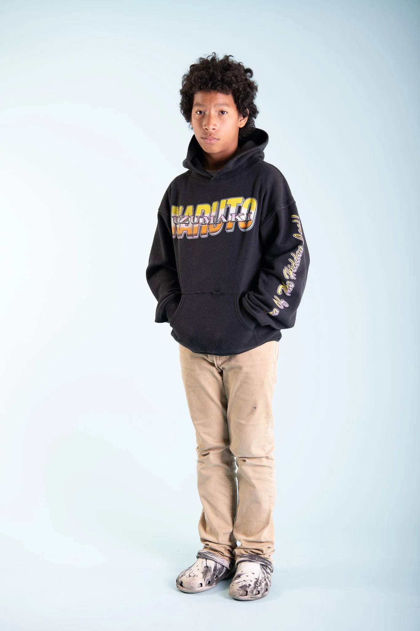 A young boy with curly hair wearing a black hoodie with colorful text and beige pants, standing with hands in pockets against a plain background.