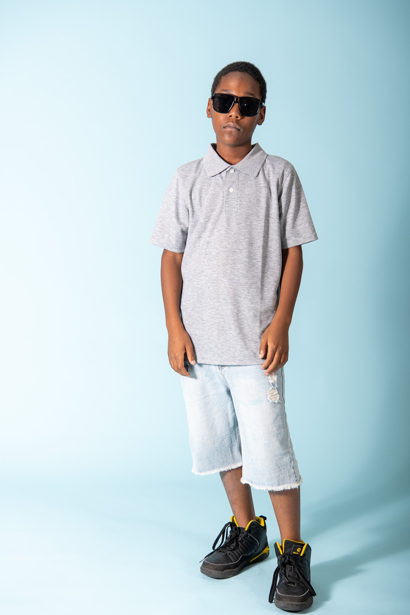 A young boy wearing black sunglasses, a gray polo shirt, ripped denim shorts, and black sneakers with yellow accents standing against a light blue background.