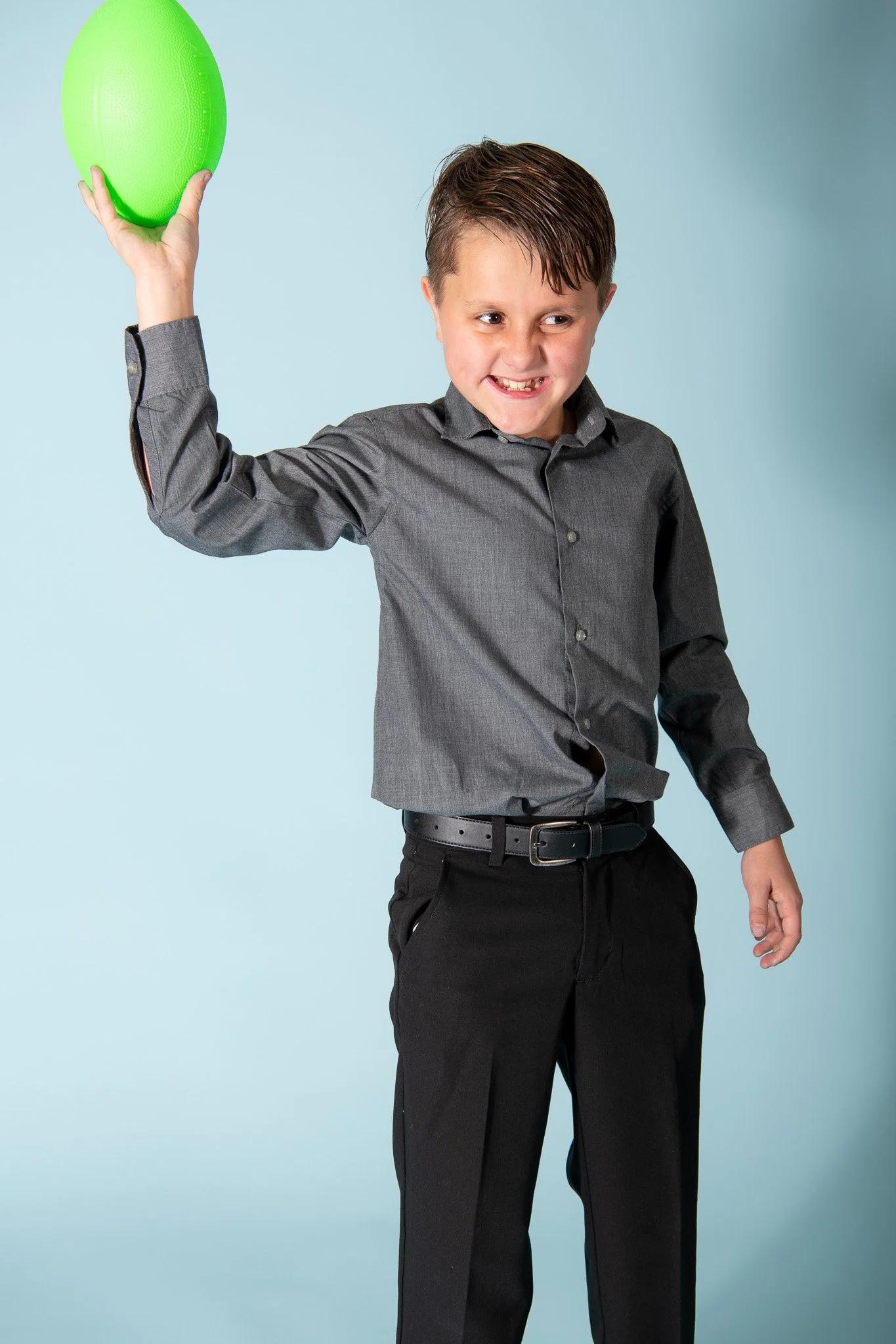 A young boy in a gray shirt and black pants holding a green ball overhead, smiling with a mischievous expression.