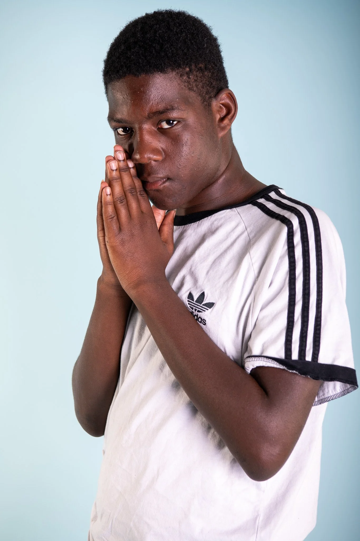 Young man with dark hair in a white Adidas T-shirt, hands pressed together near his face, looking thoughtfully at the camera, against a light blue background.