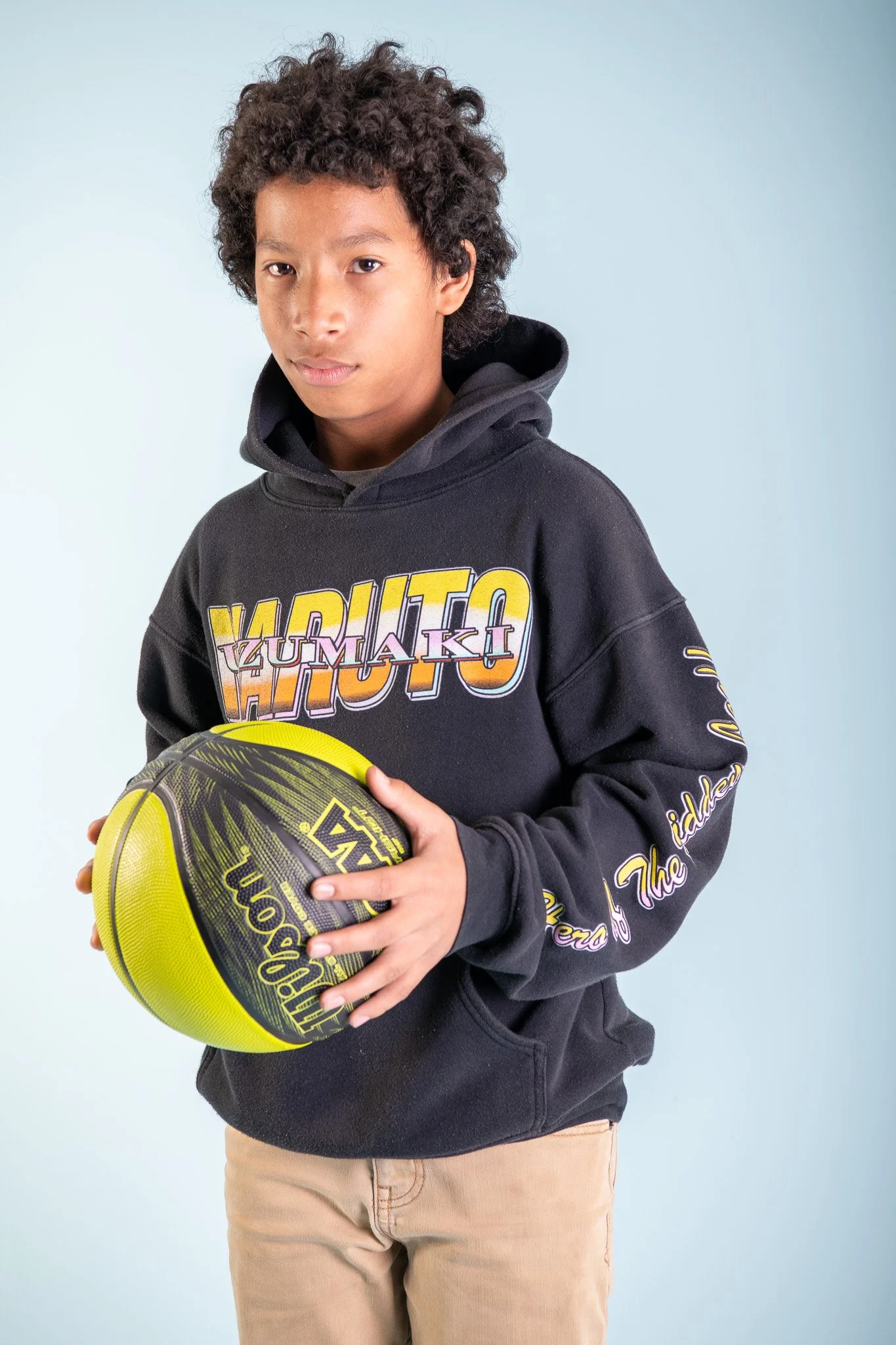 A young boy with curly hair holding a basketball, wearing a black hoodie with colorful graphic text and beige pants, posed against a light blue background.
