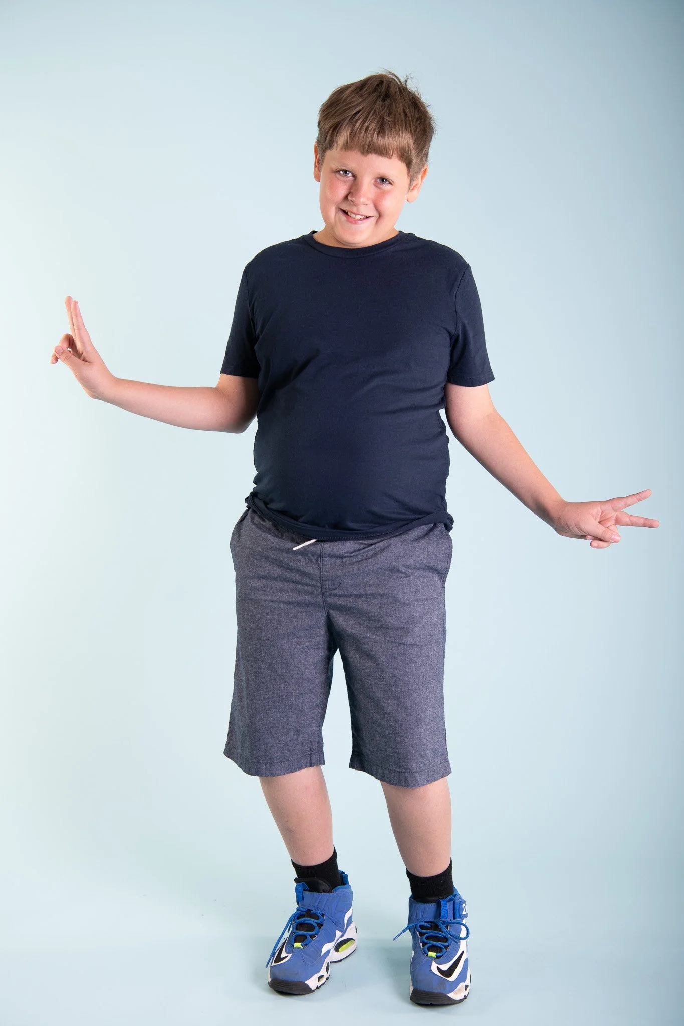 A young boy in a navy T-shirt, gray shorts, and blue sneakers posing with a happy expression and making peace signs with both hands.