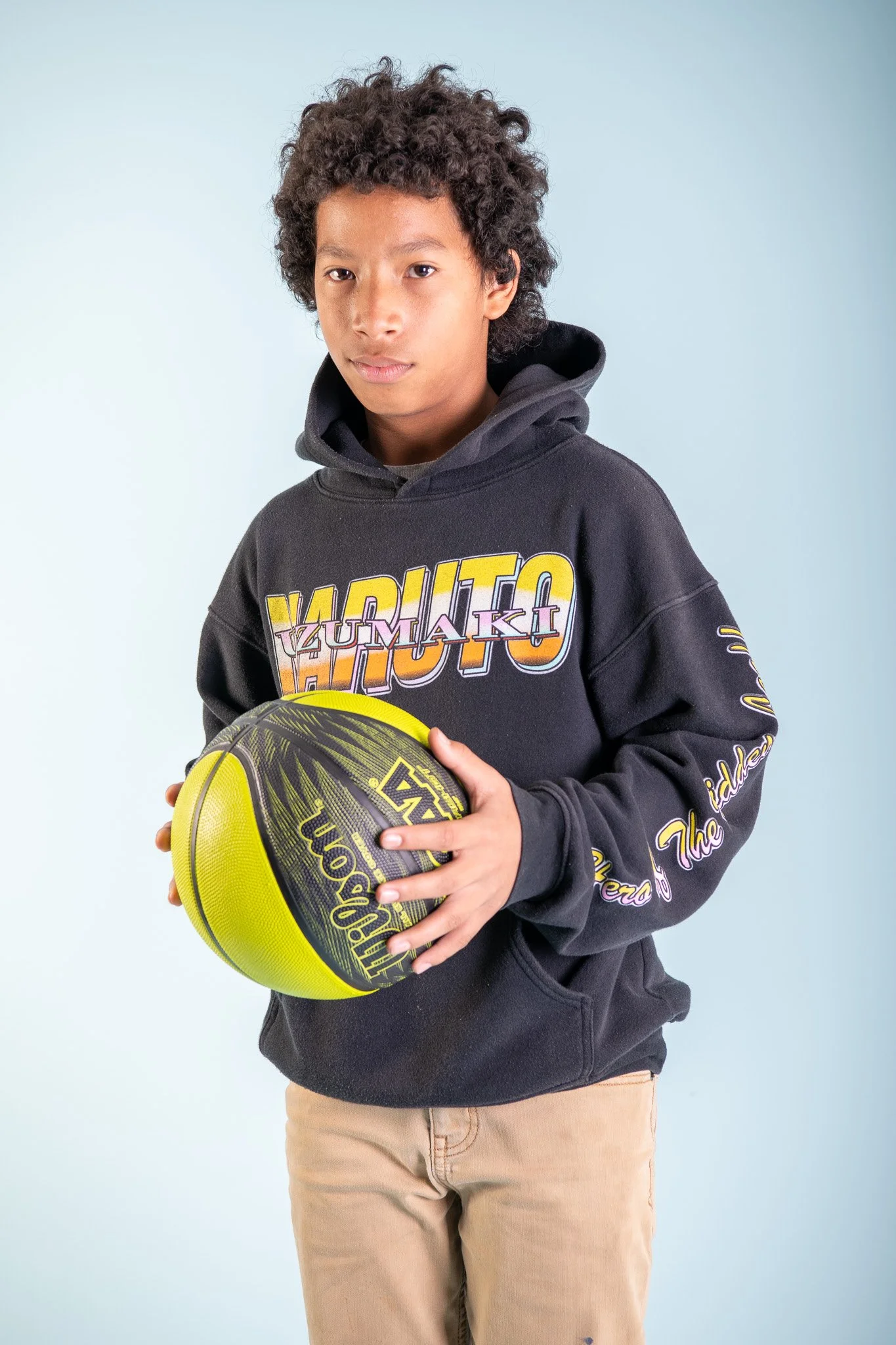 A young boy with curly hair holding a yellow and black basketball, wearing a black hoodie with colorful graphics on the front and sleeve, and khaki pants, standing against a light blue background.