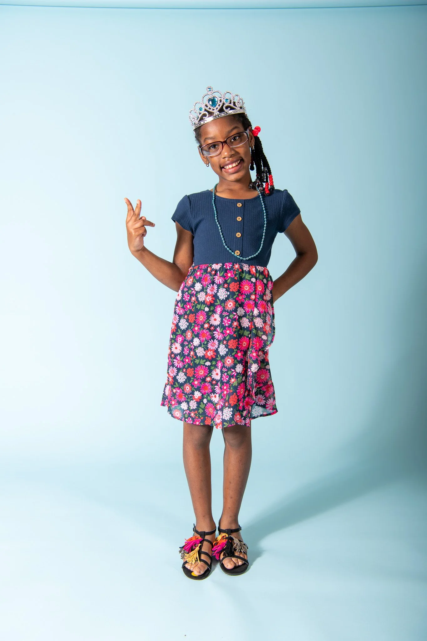 A young girl wearing a silver tiara, glasses, a blue top, a colorful floral skirt, and sandals with tassels, standing against a light blue background.