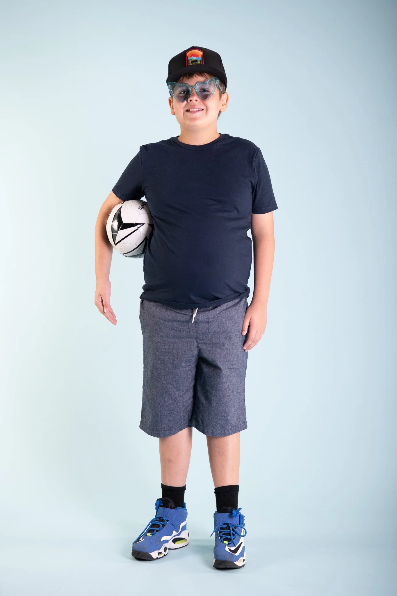A smiling young boy with glasses and a cap, holding a soccer ball under his arm, standing against a light blue background.