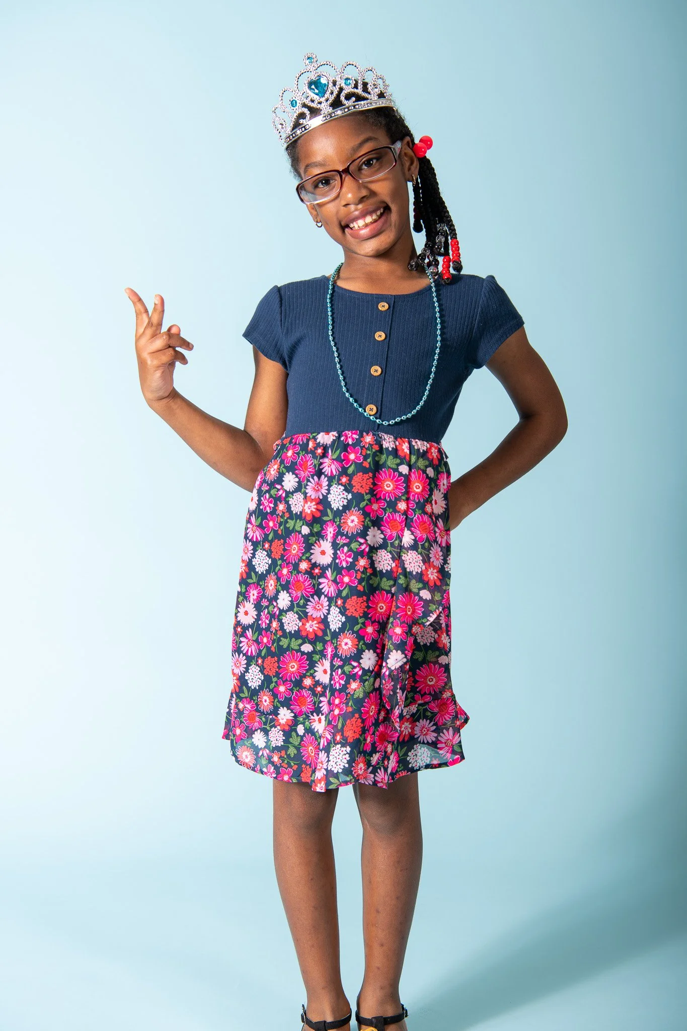 A young girl wearing glasses, a tiara, a navy blue top with gold buttons, a floral skirt, and jewelry, posing against a light blue background.