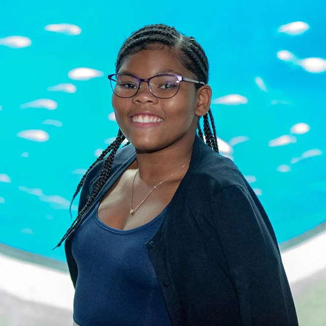 Young girl with glasses and braided hair smiling in front of an aquarium tank filled with small fish.
