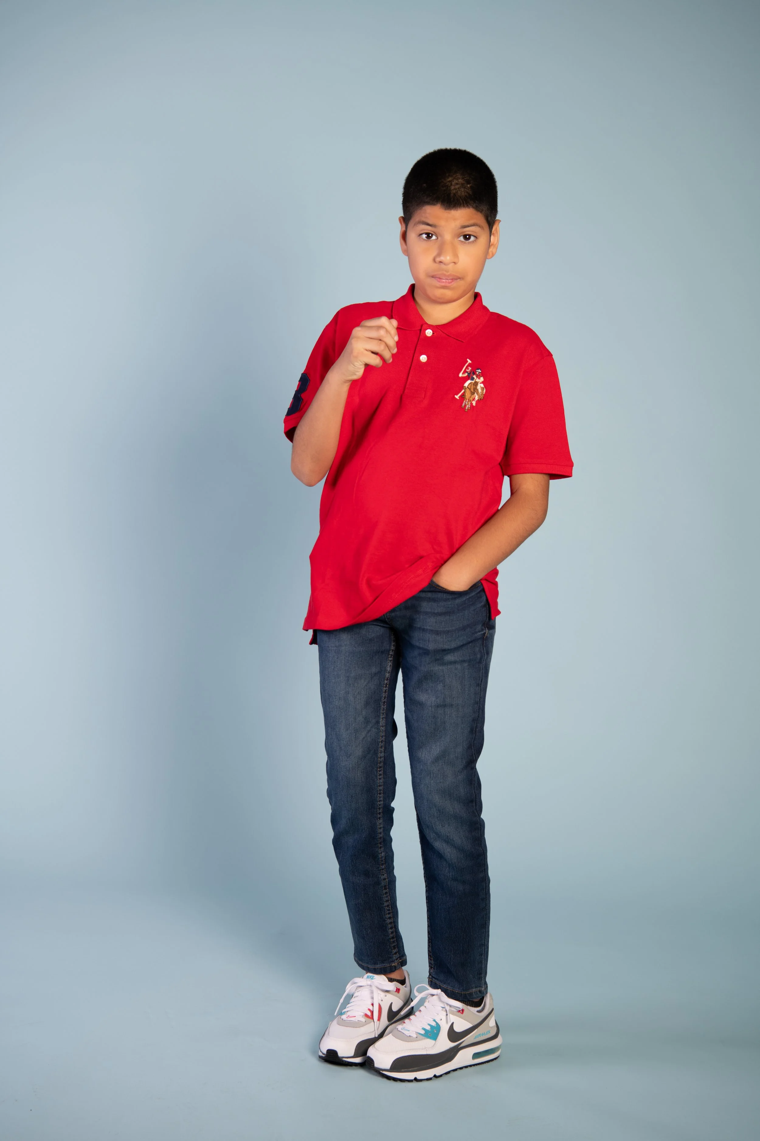 A young boy wearing a red polo shirt, jeans, and white sneakers stands against a light blue background, with one hand in his pocket and the other raised near his chest.