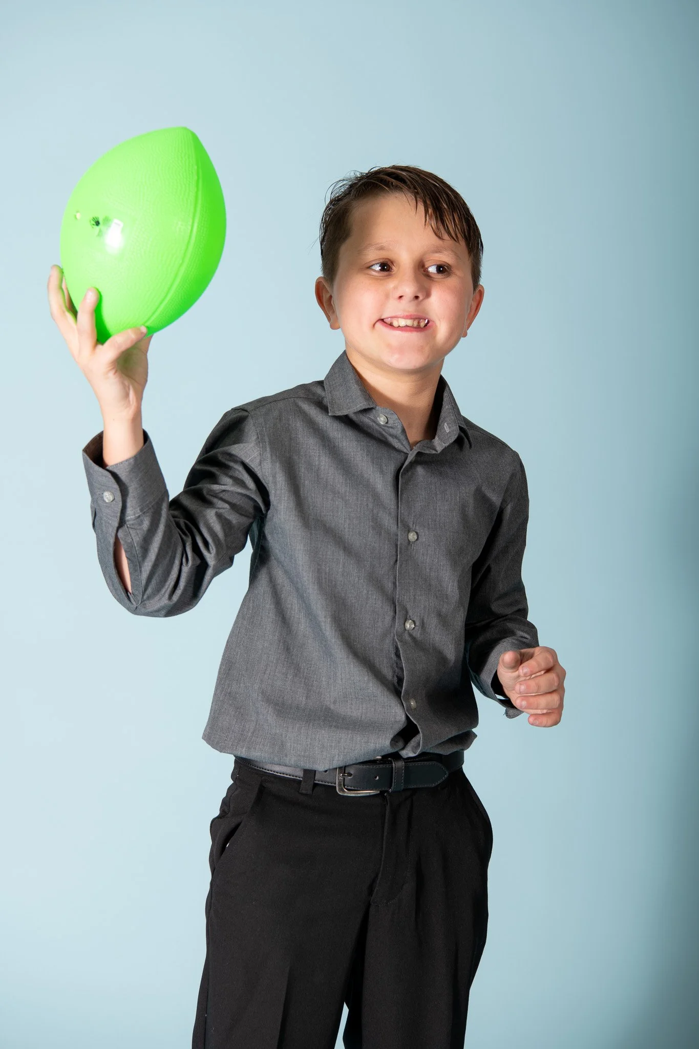 A young boy in a gray dress shirt and black pants holding a green rubber football.