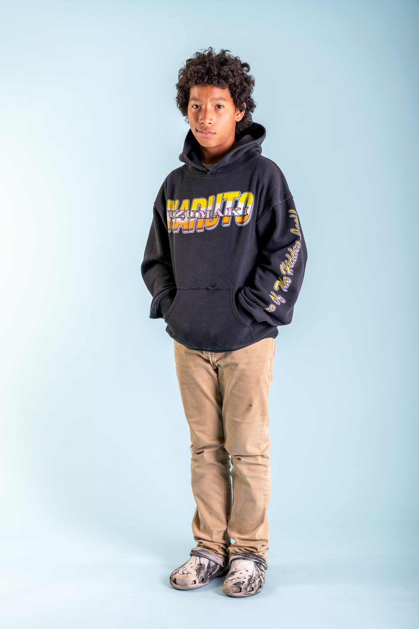 Teen boy with curly hair wearing a black Hokage hoodie, beige pants, and animal print slippers, standing against a light blue background.