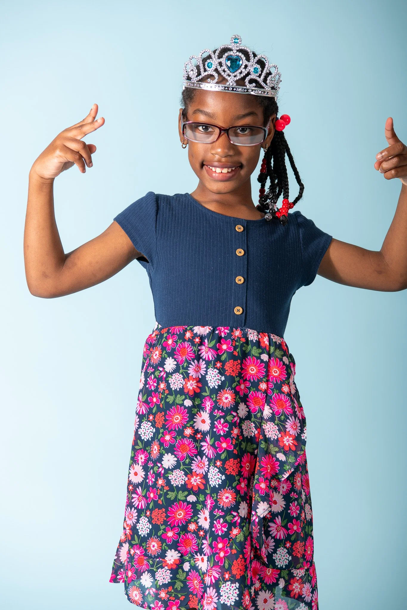 A young girl wearing glasses and a crown, flexing her arms and smiling, dressed in a blue top and a floral skirt, against a light blue background.