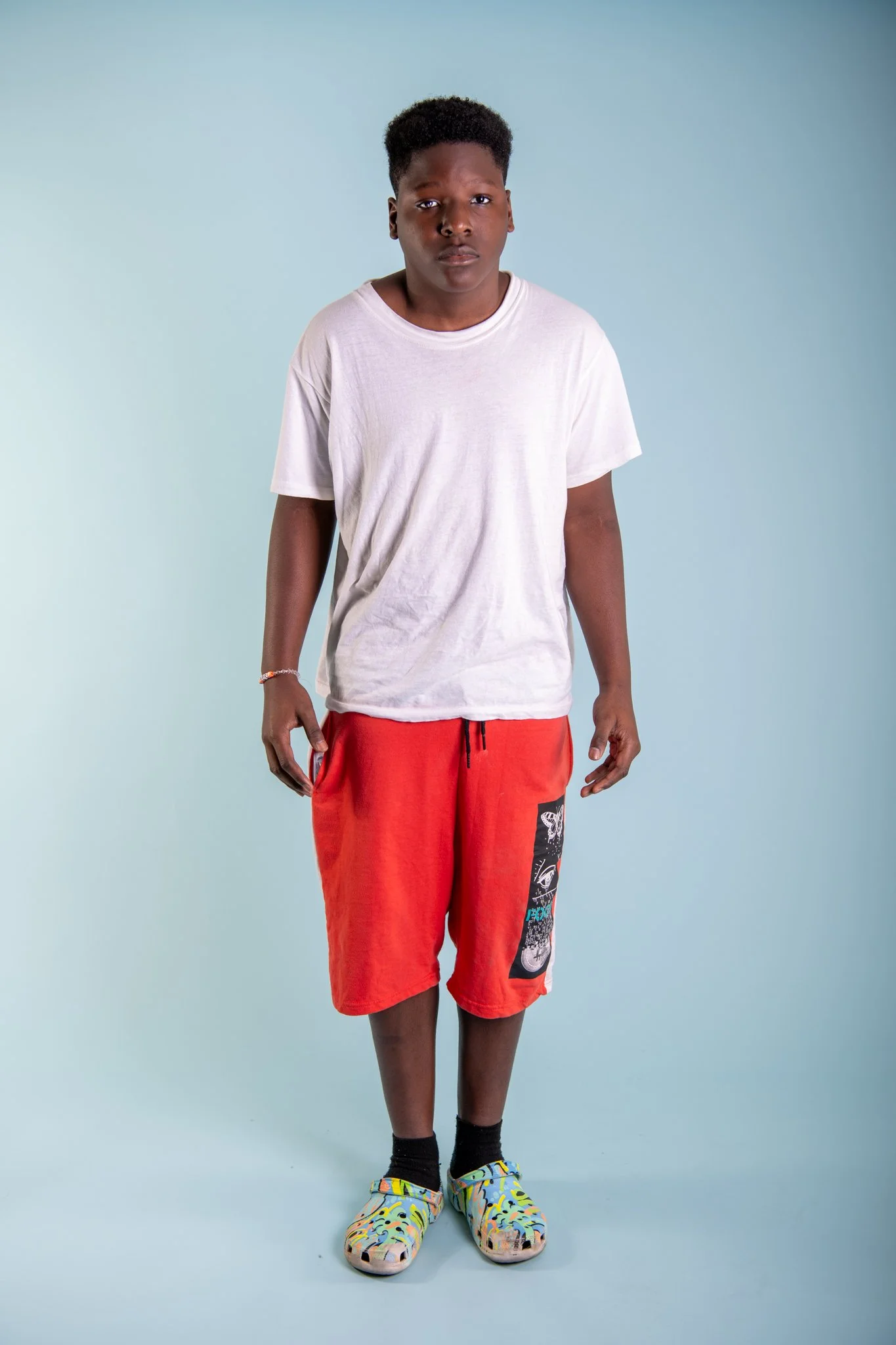 A young boy standing against a light blue background, wearing a white t-shirt, red shorts with a graphic patch, black socks, and colorful slides.