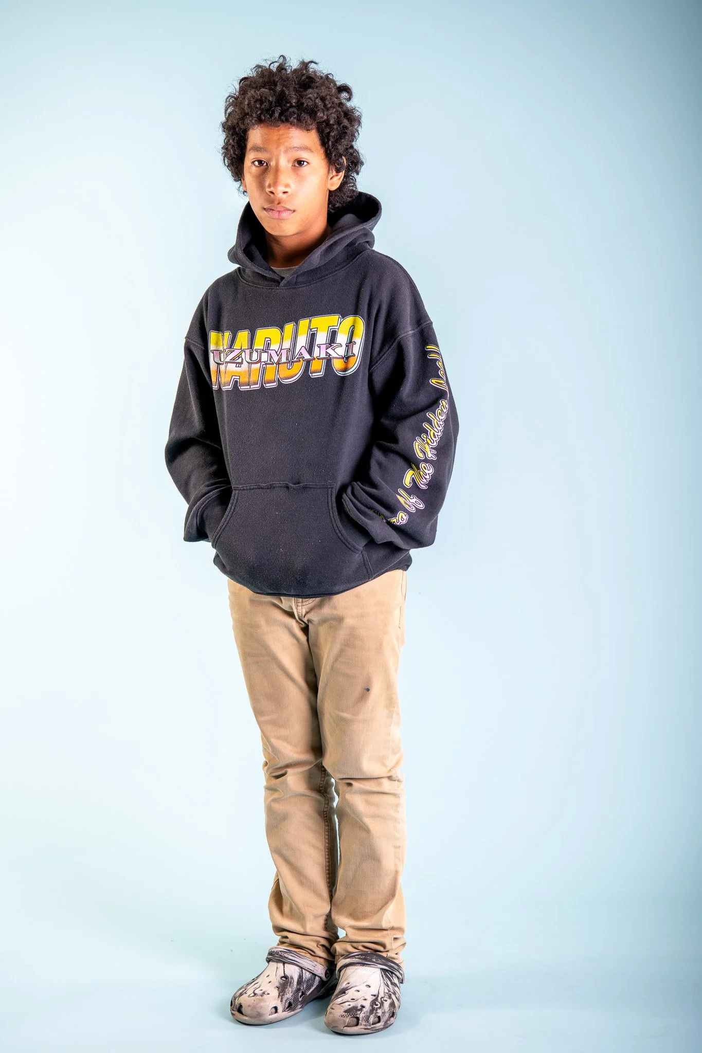 A young boy with curly hair wearing a black hoodie with colorful text, beige pants, and unique shoes, standing against a light blue background.