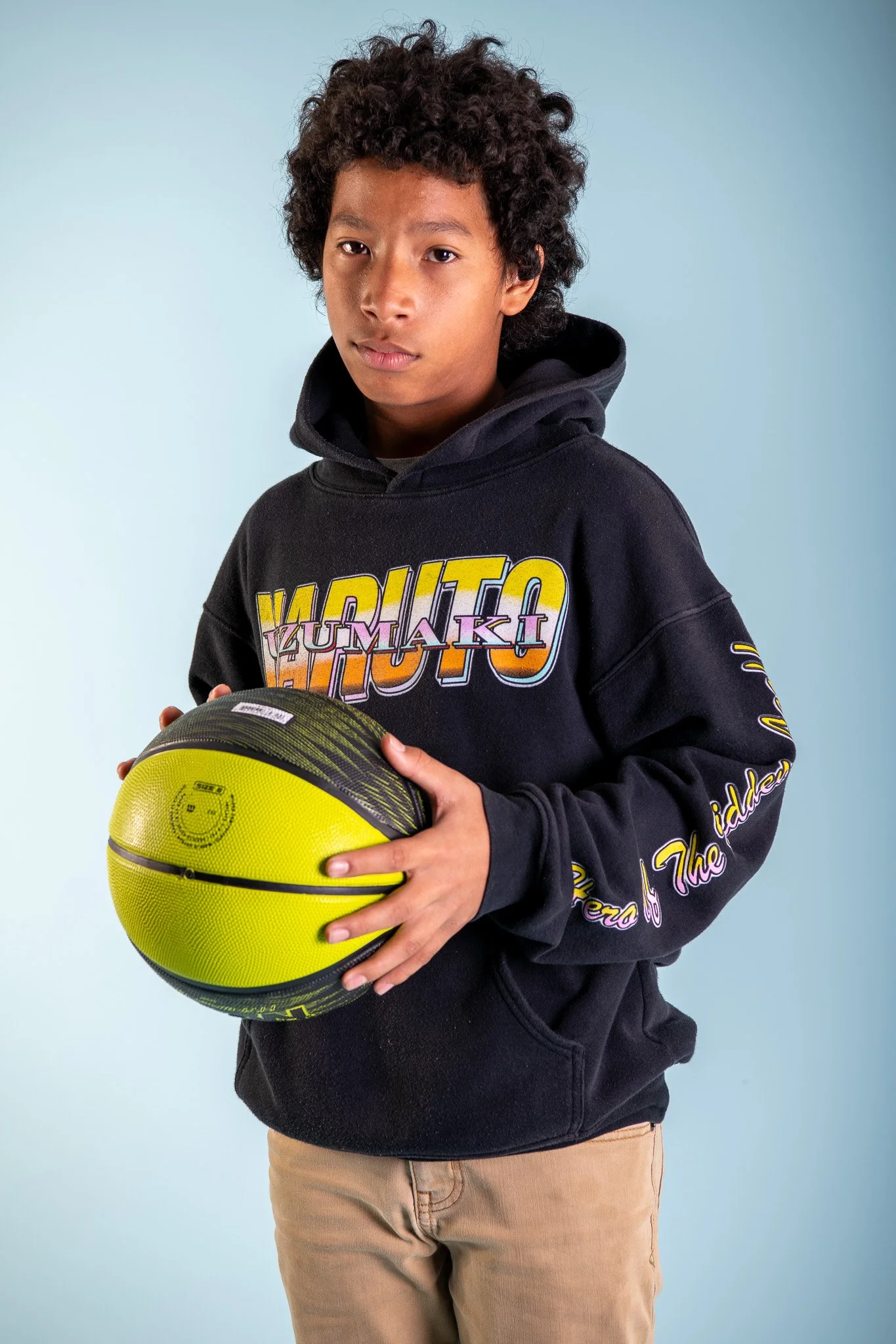 A young boy holding a yellow and black basketball, wearing a black hoodie with colorful text and beige pants, standing against a light blue background.