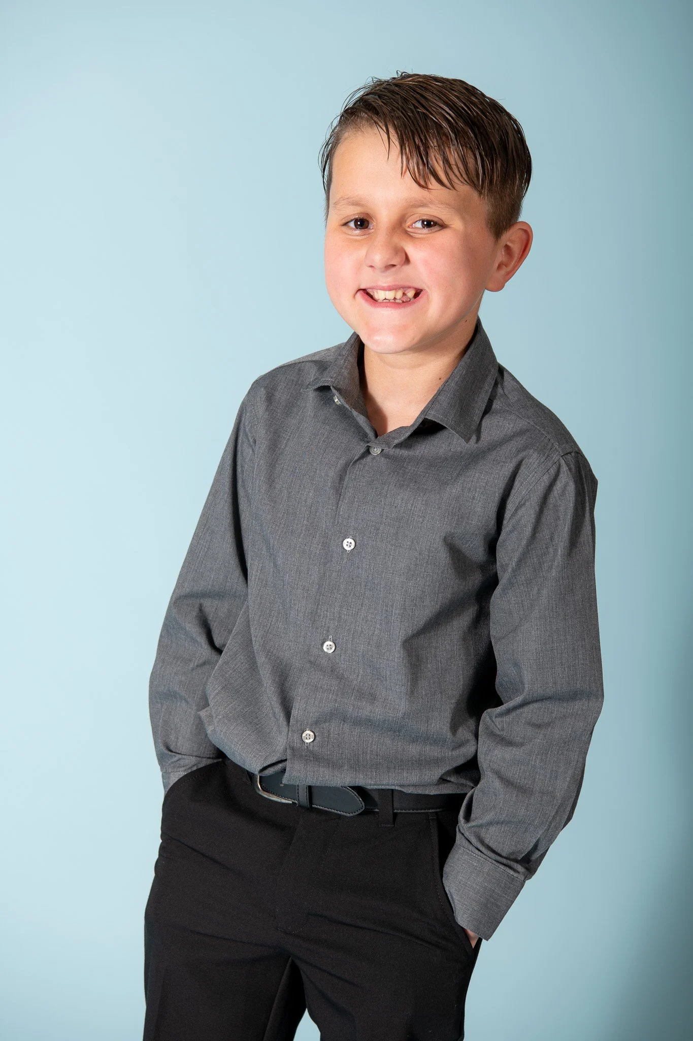 A young boy smiling, wearing a gray button-up shirt and black pants, with his hands in his pockets, standing against a light blue background.