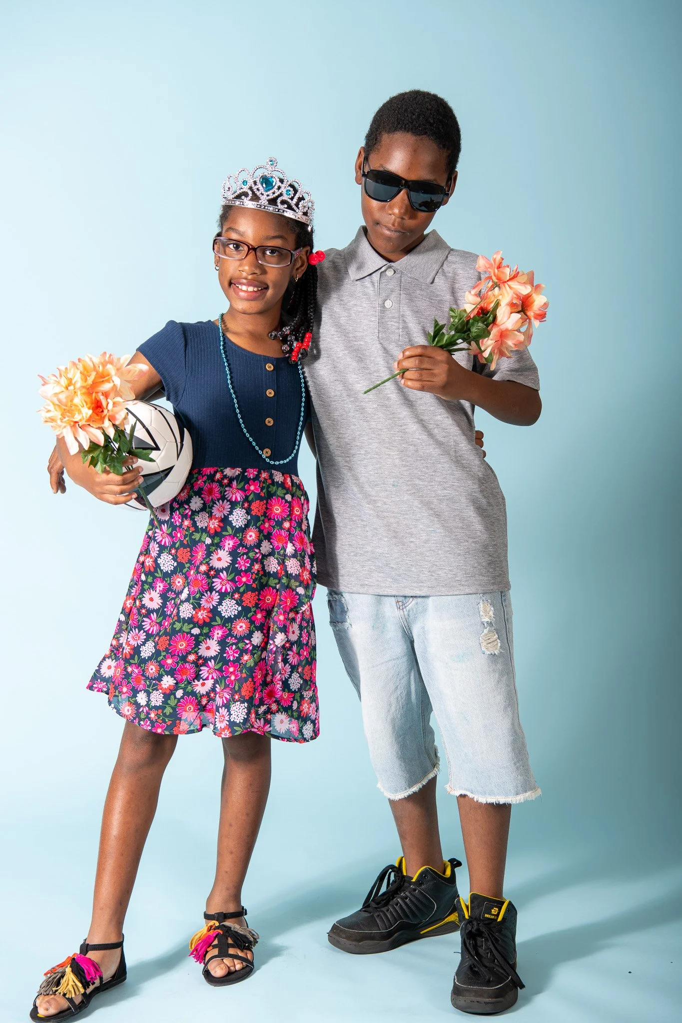 A young girl wearing glasses and a crown, holding flowers and a soccer ball, standing next to a boy in sunglasses and casual clothes, holding flowers, both smiling against a light blue background.