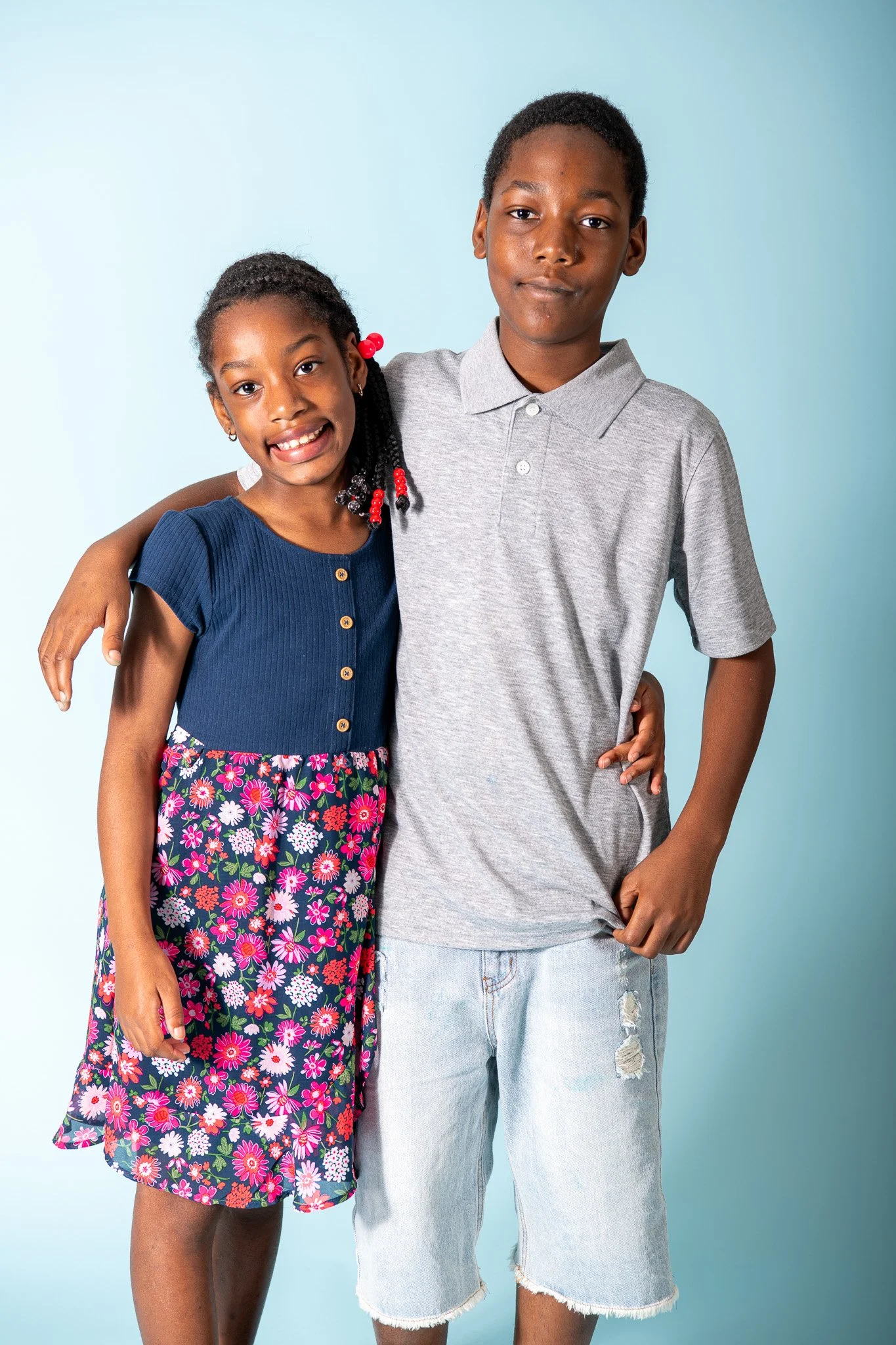 A young girl and boy stand together with their arms around each other, smiling at the camera against a light blue background.