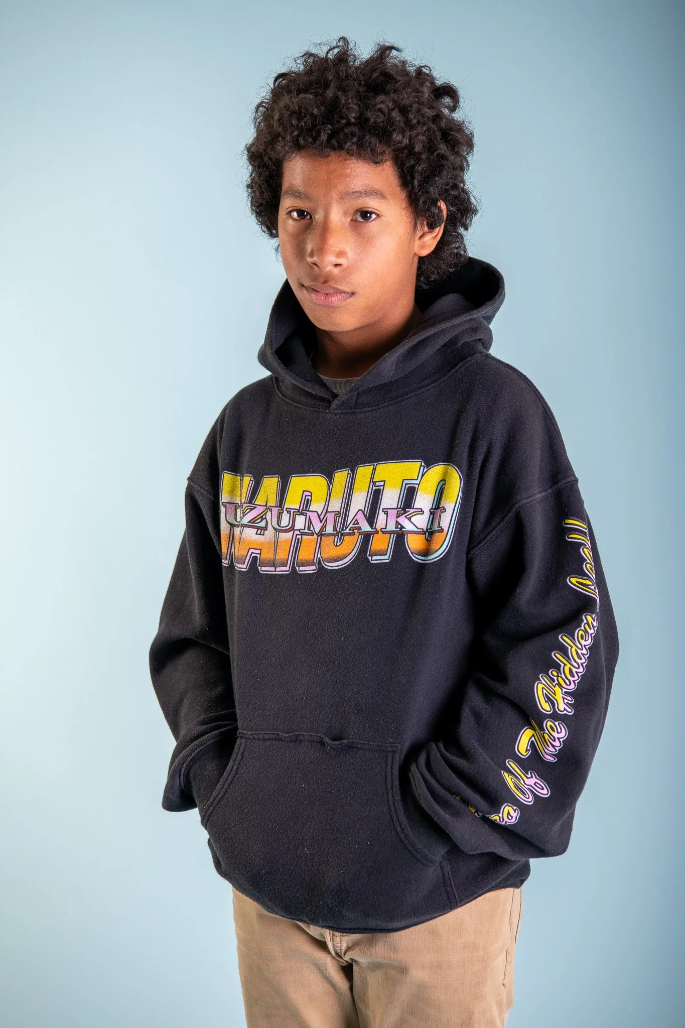 Young boy with curly hair wearing a black hoodie with colorful text and beige pants, standing against a light blue background.