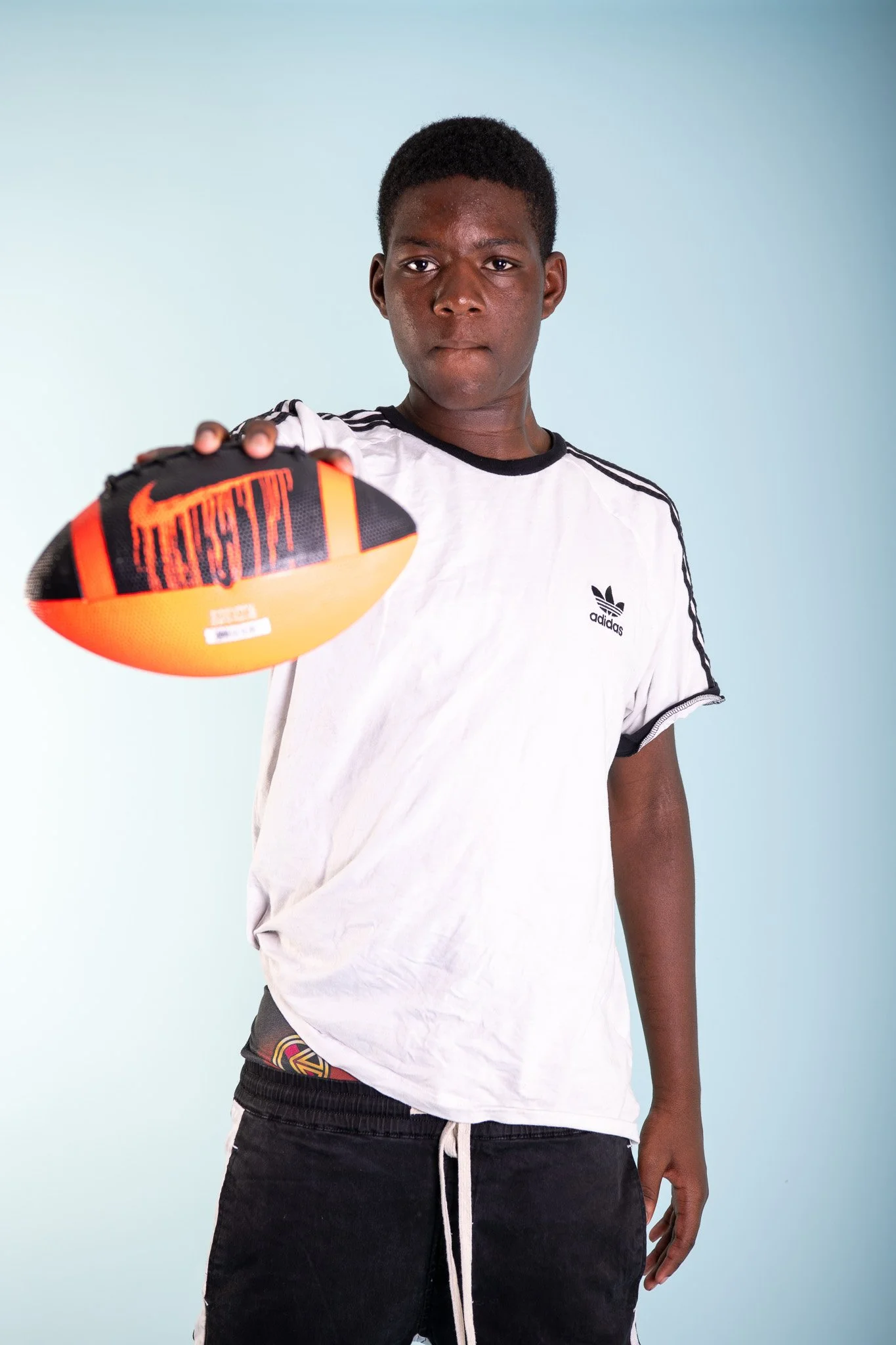 A young man wearing a white Adidas shirt and black shorts holding a football toward the camera.