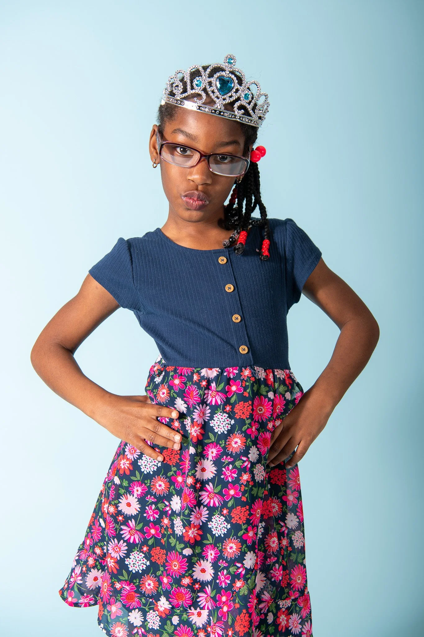A young girl wearing a crown, glasses, a blue top, and a floral skirt, standing with her hands on her hips against a light blue background.