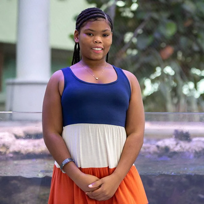 Young girl standing outdoors in front of a water fountain, wearing a color-blocked dress with a blue top, white middle, and orange skirt, smiling at the camera.