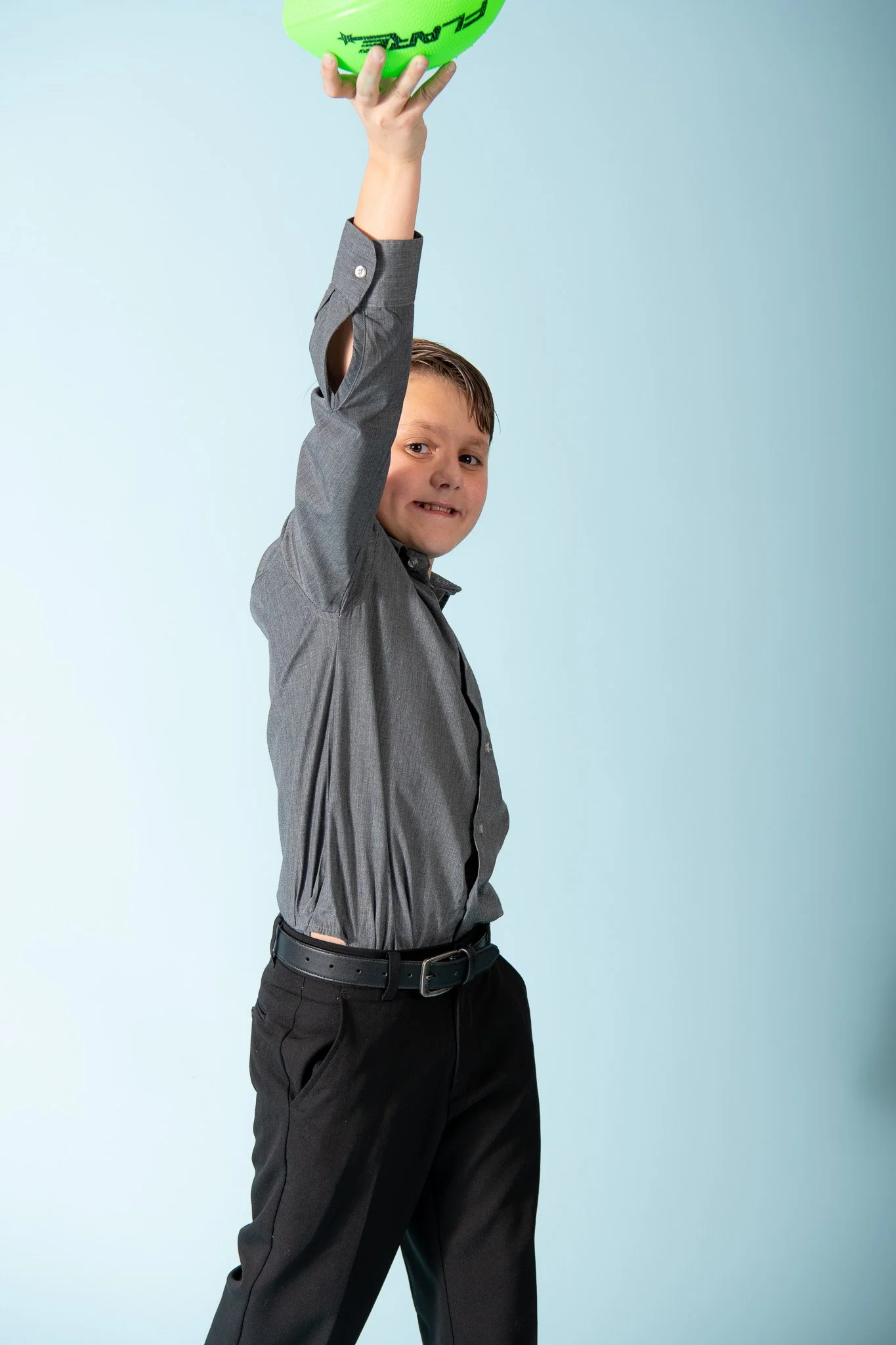 A young boy dressed in a gray shirt and black pants holding a green football above his head, smiling at the camera.