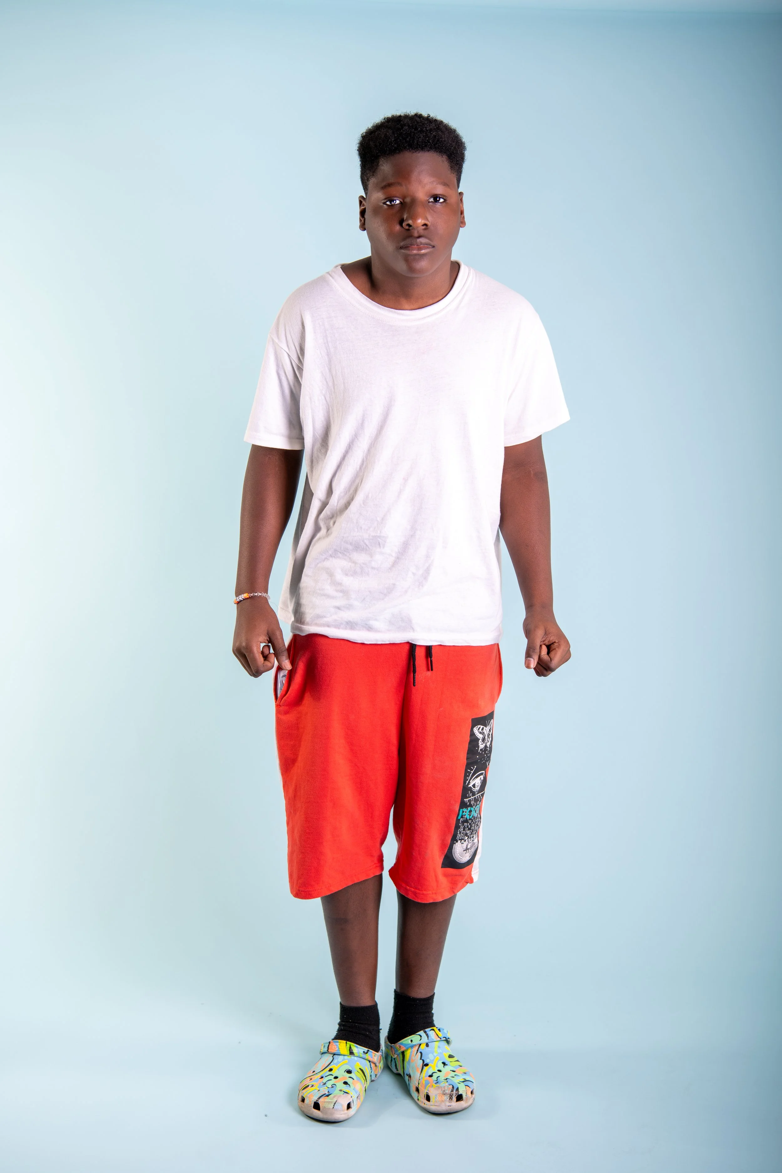 A young man standing against a light blue background, wearing a white t-shirt, red shorts, black socks, and colorful slide sandals.