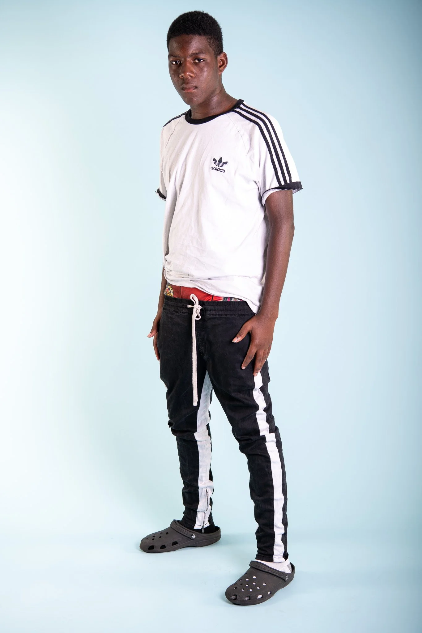 A young man standing in front of a light blue background, wearing a white Adidas t-shirt, black track pants with white stripes, and gray Crocs.