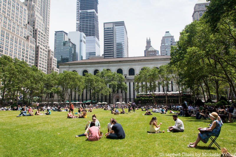 People relaxing and enjoying a sunny day in a park with lush green grass and trees, surrounded by tall city buildings.