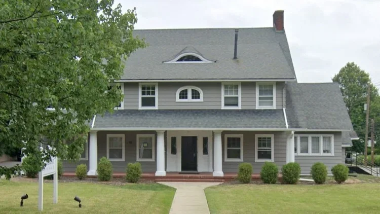 Front view of a two-story gray house where the Therapists of New York Monclair office is located, with white trim, a covered porch, and a front yard with grass, bushes, and a sidewalk.