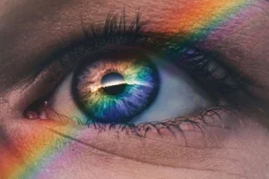 Close-up of a human eye with rainbow reflections and a rainbow light streak across the eye and surrounding skin.