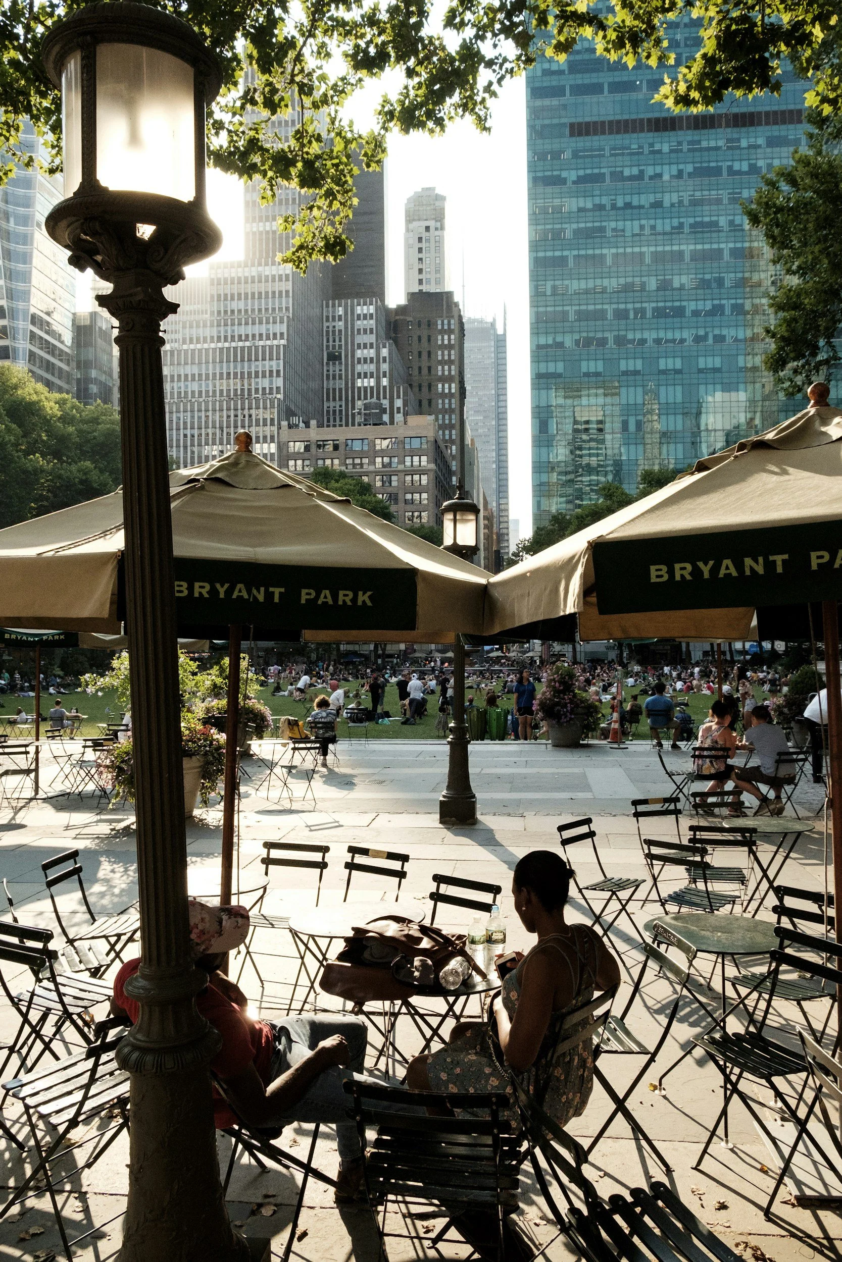 People sitting at tables and chairs in Bryant Park on a sunny day, with tall skyscrapers in the background and umbrellas providing shade.